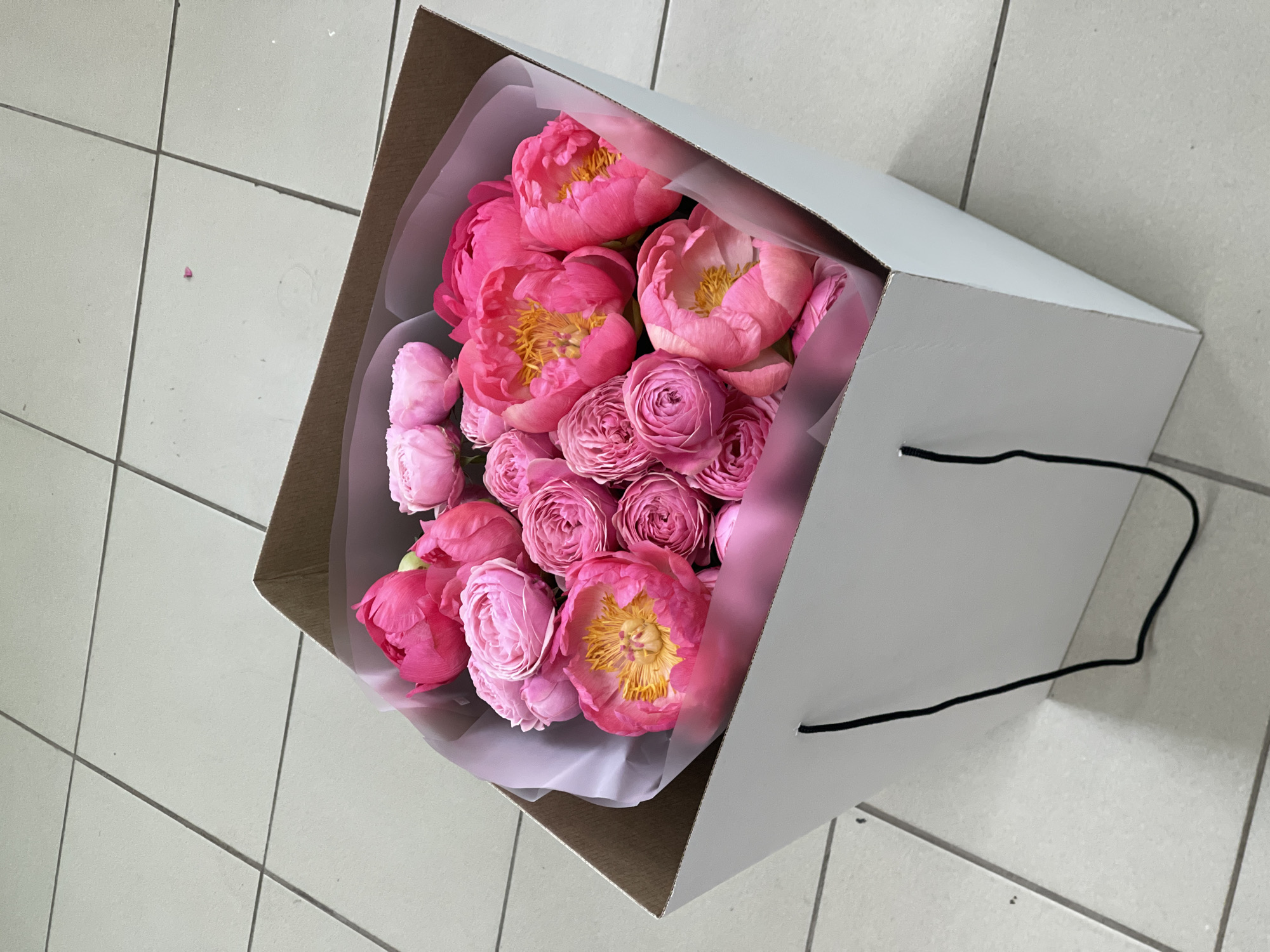 A bird's-eye-view image shows a white box filled with pink flowers, sitting on a floor made of light gray square tiles. The box has black string handles and appears to hold a bouquet of roses and peonies wrapped in light-colored tissue paper. The flowers are densely packed within the box, exhibiting various shades of pink with some having yellow centers. The tiled floor provides a neutral background with grid lines between the tiles. The location of Izhevsk, Russia is not evident from the image.