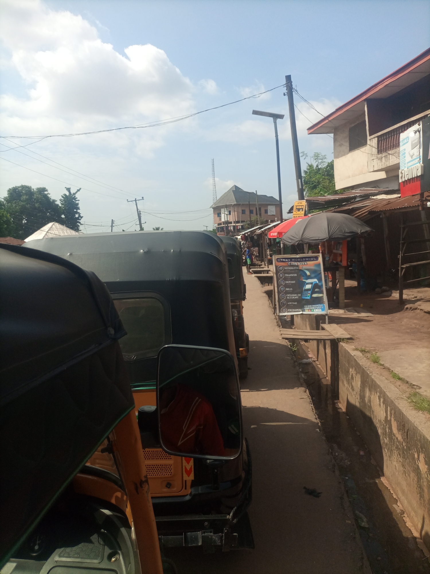 This media file depicts a street scene in Nkpor, Nigeria, likely during the daytime under a partly cloudy sky. The foreground features the interior of a vehicle, possibly a three-wheeled auto-rickshaw, with a prominent side mirror reflecting a person wearing a red top with white stripes. The view is directed down a street lined with buildings and market stalls on the right side.
Several auto-rickshaws are parked in a queue, suggesting a busy area or a waiting point. The market stalls are constructed with corrugated metal roofs and wooden planks, typical of informal marketplaces in the region. A prominent sign in front of one stall advertises "TEEZY WORLDWIDE Cash bank" with services like withdrawal, transfer, deposit, bills, opening of account, and airtime. There's also a phone number visible on the sign.
Further down the street, more buildings and structures are visible, including what appears to be a cell tower in the distance. A concrete drainage channel runs along the side of the street, containing some water and debris. The overall impression is of a bustling urban environment with everyday economic activities taking place. The lighting suggests it's daytime, and the clouds indicate a mix of sun and shade.