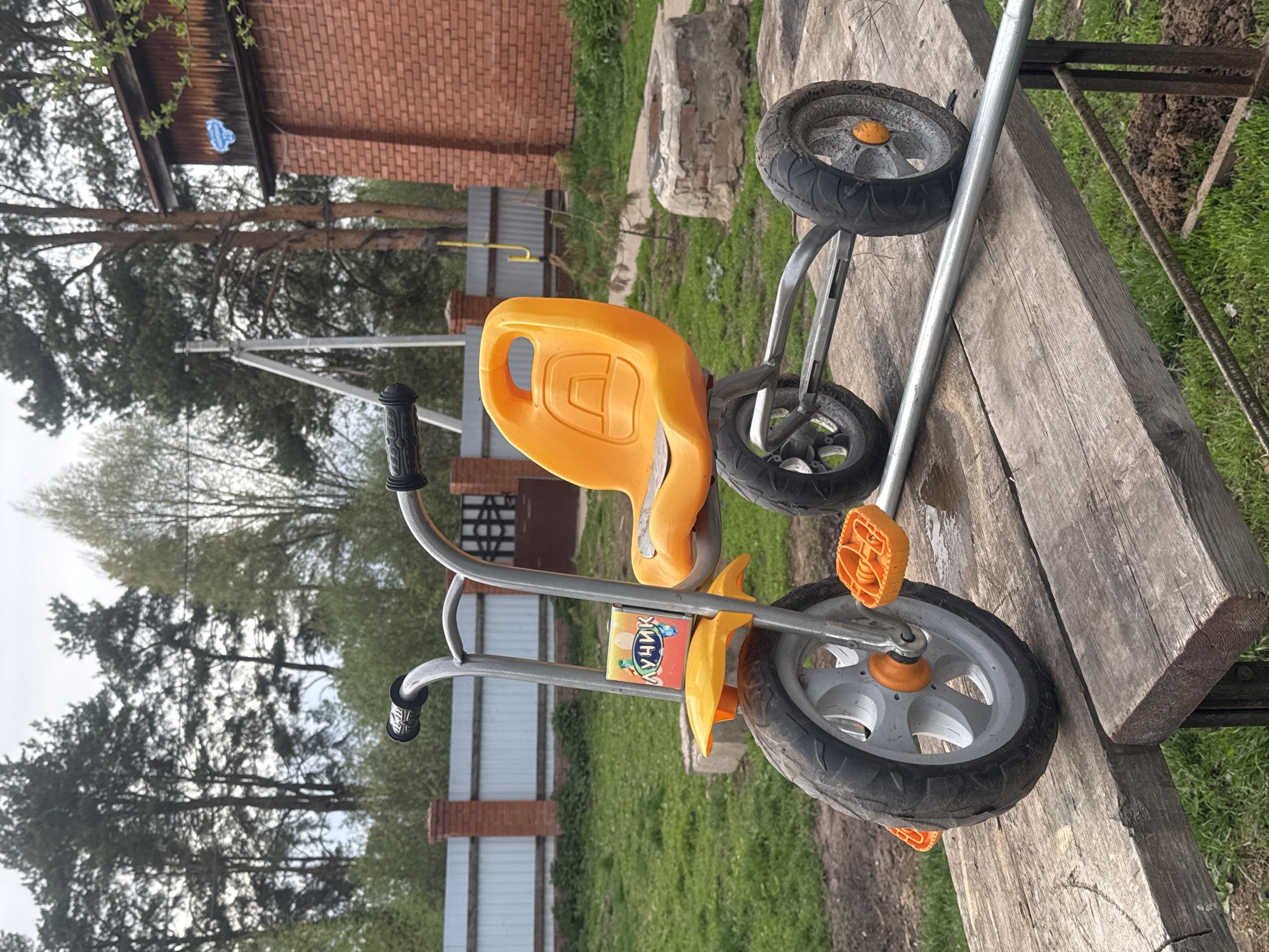 A child's tricycle rests atop a weathered wooden surface, potentially a bench or table. The tricycle has an orange seat, an inscription in Cyrillic script on the frame, and grey wheels. The surroundings include grass, trees, and a brick structure, possibly a building or fence. The image appears to be taken outdoors in a residential or park area. Based on the information provided, the location is Rusalevka, Russia.