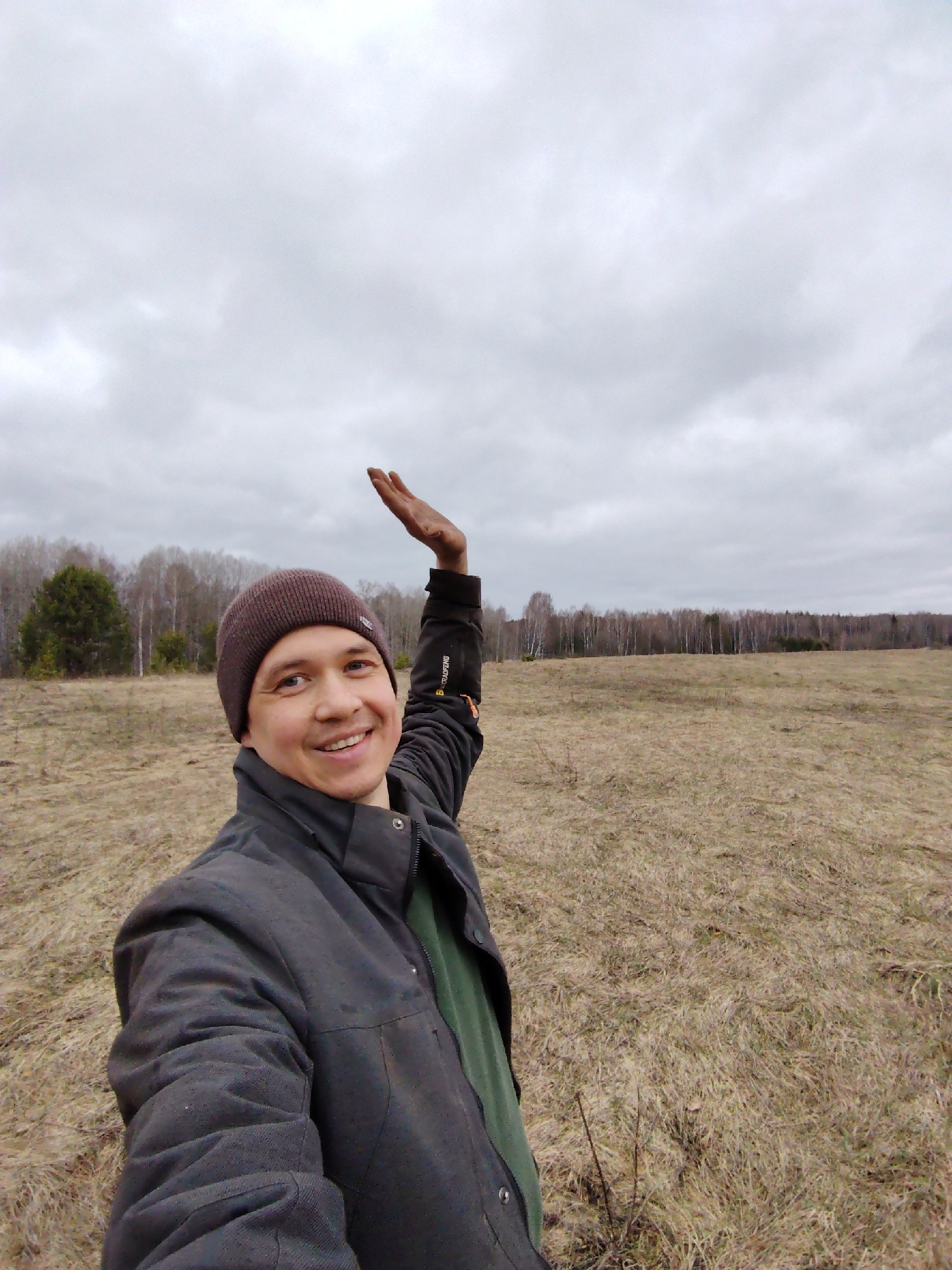 An outdoor selfie captures a smiling man on an overcast day in Markovskii, Russia. The man, positioned in the foreground, grins directly at the camera. He wears a dark grey or black jacket, a green shirt, and a dark brown knit beanie hat. His right arm is raised with his hand held upwards, displaying some dirt or grime on his palm.
The background reveals a wide, open field covered in dry, light brown and yellowish grass, suggesting either early spring or late autumn. A distant forest line stretches across the horizon, primarily consisting of bare deciduous trees, with a cluster of darker evergreen trees visible on the left side. The sky is entirely overcast, dominated by a uniform grey cloud cover. The ground immediately around the man is uneven, with patches of dry vegetation and some visible dark earth. Text resembling "B.TRADEPORT" is faintly visible on the man's jacket sleeve.