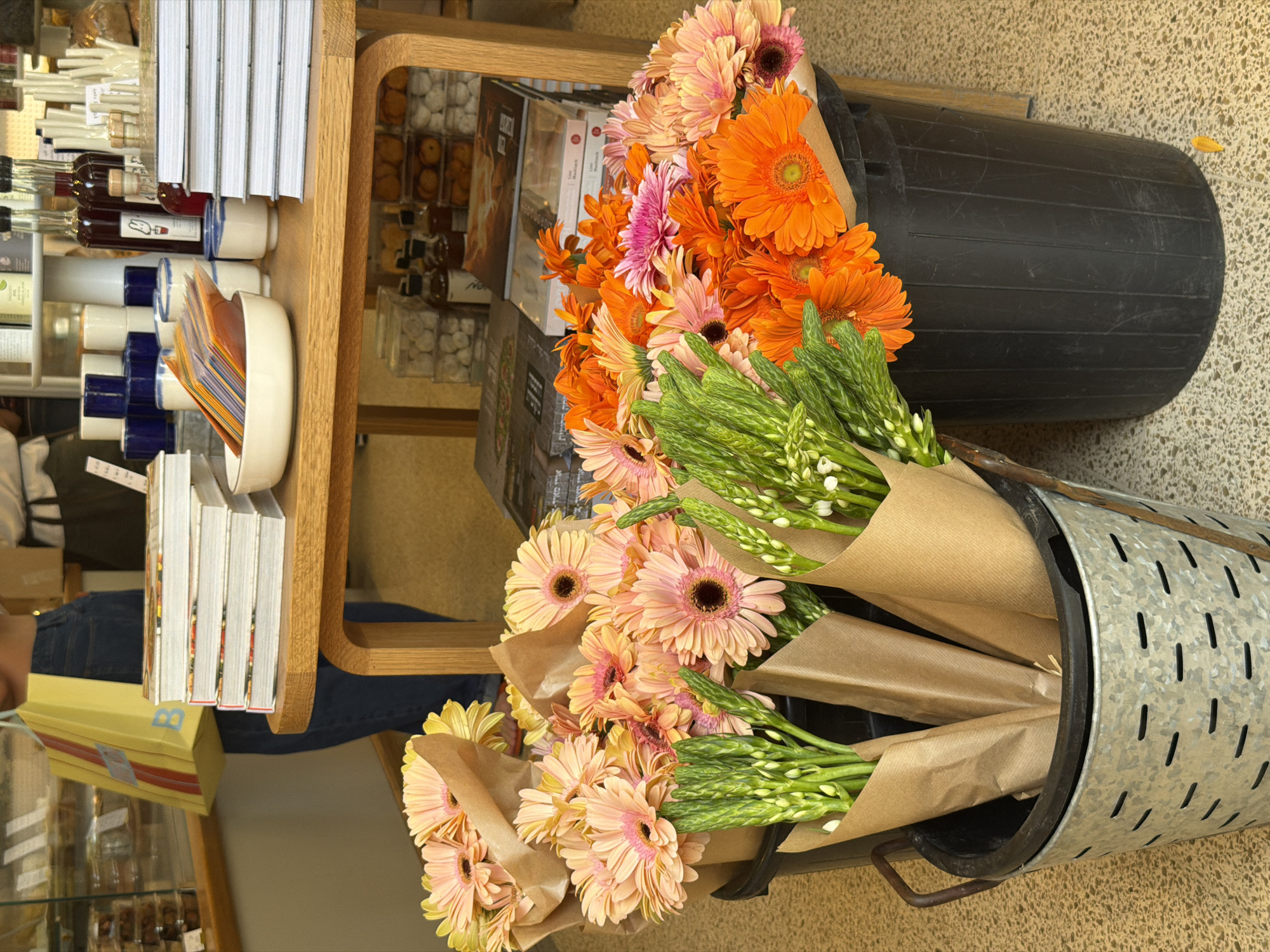 The image depicts a collection of cut flowers displayed within a shop. The flowers consist of gerbera daisies in shades of peach, orange, and pink, along with stalks of green asparagus flowers. The flowers are arranged in bouquets, wrapped in brown paper. Some of the bouquets are placed in a perforated metal bucket on its side. Other flowers are gathered in a tall, black, round bin. Adjacent to the flowers is a wooden shelving unit displaying various products, including books, bottles, and packaged goods. The location is a shop in Tel-Aviv, Israel.