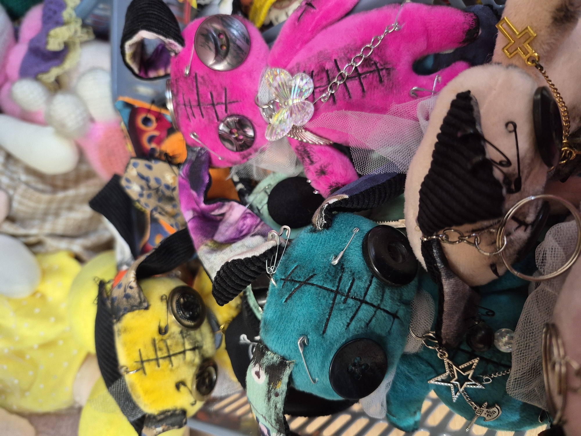 The image displays a collection of handmade stuffed toys, possibly in a craft market or workshop setting in the city of Krasnyy Klyuch, Russia. The toys are eclectic in design, with a focus on a "creepy-cute" aesthetic.
One prominent toy is bright pink, with large black buttons for eyes and stitched details. It is adorned with a delicate iridescent butterfly charm and a silver chain. Another toy, positioned to the left and slightly behind the pink one, is teal-colored, also featuring button eyes and stitched markings. It has safety pins attached to its body. A yellow toy, partially visible in the upper left, shares similar button eyes and stitched features, with a black and patterned fabric headpiece. Other toys are visible in the background, some appearing to be made of pastel-colored yarn or fuzzy material.
The toys are arranged in what appears to be a basket or container, with various fabric scraps and embellishments scattered around them. The lighting suggests an indoor setting, possibly with natural light filtering in. The overall impression is one of artisanal crafting, with a focus on unique, characterful creations.
