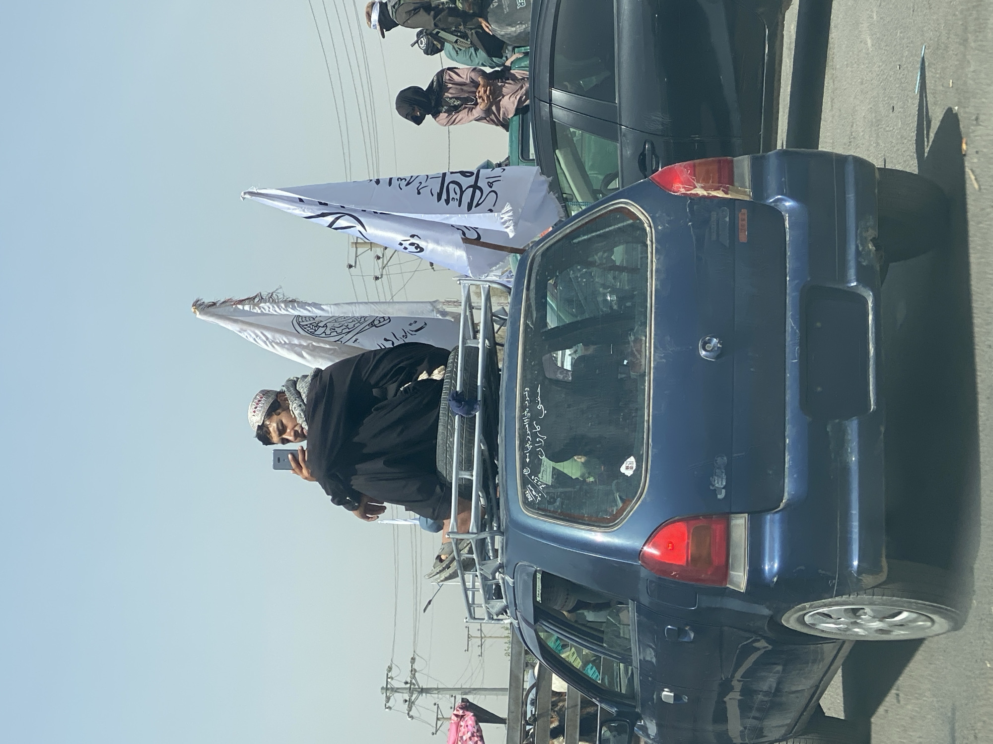 In a street scene in Kandahar, Afghanistan, on a clear day, a man wearing a white and patterned cap and a dark robe is seated on the roof rack of a blue car, holding a mobile phone. He appears to be taking a photo or video. Two white flags with Arabic script are visible behind him, mounted on poles. The car's rear window has some Arabic writing on it. Other vehicles and people are present in the background, including a woman in a light brown headscarf and a person in a black head covering in another vehicle behind the blue car. Power lines and poles are visible in the distance. The overall atmosphere suggests a procession or gathering.