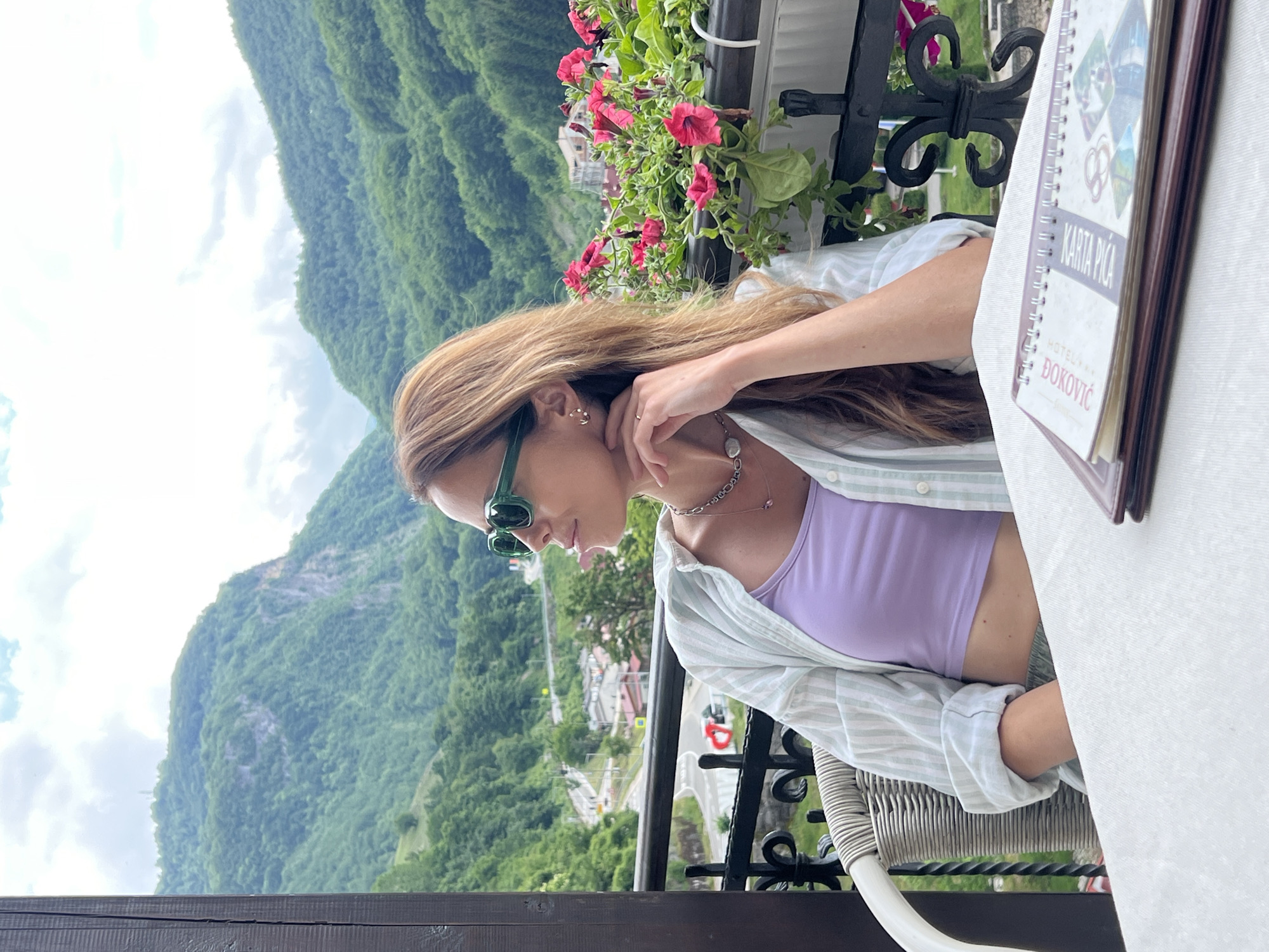 This outdoor daytime image features a woman seated on a balcony, enjoying a panoramic view of a mountainous landscape, identified as Šavnik, Montenegro. The woman has long, light brown hair and wears green-framed sunglasses, a light purple crop top, and an open, light-colored striped button-down shirt. She is adorned with a delicate necklace and earrings, and her hand rests near her neck.
She is positioned at a table with a white tablecloth, and a portion of a white wicker-style chair is visible. An open menu, featuring the text "KARTA PIĆA" (Drink Menu) and "HOTEL *** ĐOKOVIĆ," lies on the table, indicating a restaurant or cafe setting. A decorative black metal railing, topped with a flower box bursting with pink petunias, separates the balcony from the stunning view. Below, a valley stretches out, revealing a winding road and glimpses of buildings amidst the lush greenery.
The background is dominated by majestic, green-clad mountains under a bright, partly cloudy sky, suggesting pleasant weather. The overall atmosphere is relaxed and scenic, with the woman seemingly lost in contemplation or enjoying the picturesque surroundings. The location clearly offers breathtaking natural beauty, typical of a mountainous region.