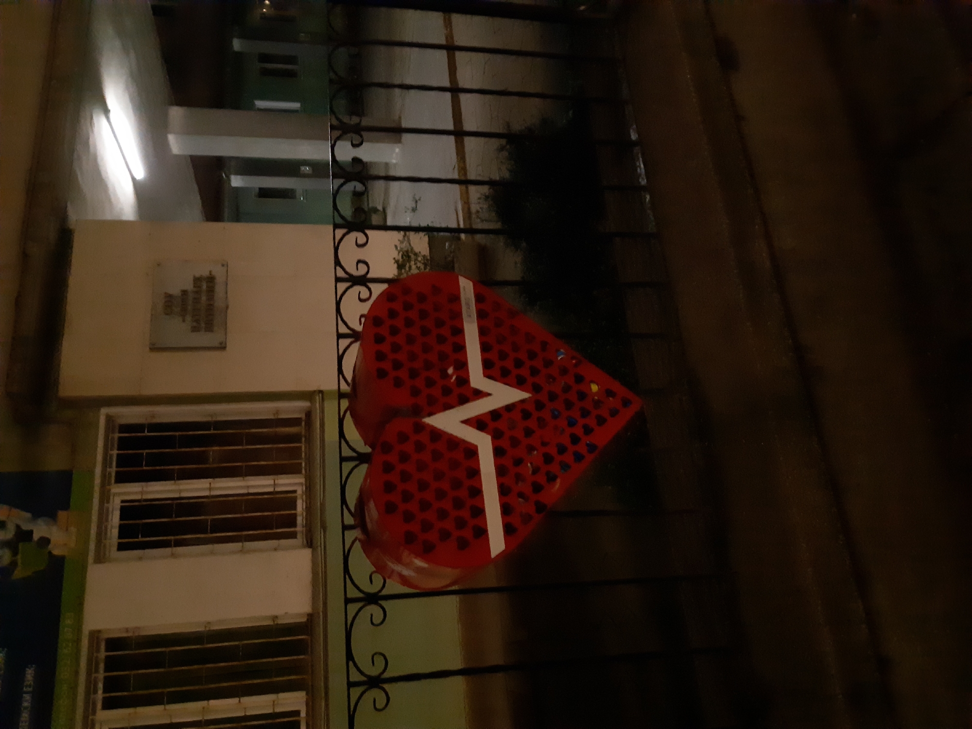 A heart-shaped object with a white zigzag line is attached to a wrought-iron fence. The object is red and appears to be constructed from multiple heart shapes. The fence is in front of a building with barred windows and a plaque. The plaque reads "ODZ (children's complex) 'Radost' Region 'Izgrev.'" This suggests the location is likely in Plovdiv, Bulgaria, given that Izgrev is a region of the city. The scene appears to be at night.