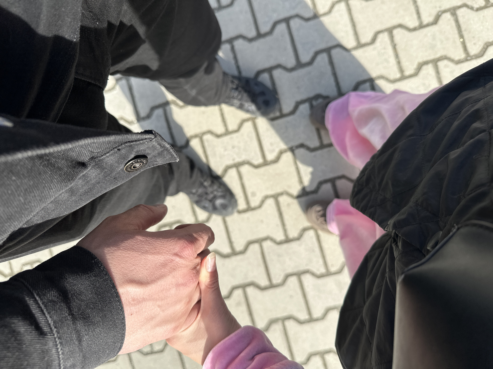 A photograph depicts two individuals standing on a paved walkway composed of interlocking concrete pavers. The pavers are light gray and arranged in a repeating pattern. The sun casts shadows, indicating a daylight setting.
The person on the left is wearing a black denim jacket with a visible button closure. The visible skin on the left subject is light-skinned. They are holding hands with another individual on the right, who is wearing a pink garment, possibly a jacket or sweatshirt. The person on the right is wearing a black quilted jacket.
The footwear of both individuals is visible. The person on the left is wearing dark-colored shoes with a patterned design, and the person on the right is wearing gray shoes. The focus of the image is on the holding of hands, suggesting a close relationship.
Based on the location metadata and the prevalence of concrete paver walkways in urban areas, the setting is likely a public space in Yekaterinburg, Russia. The specific location cannot be determined from the visual data.