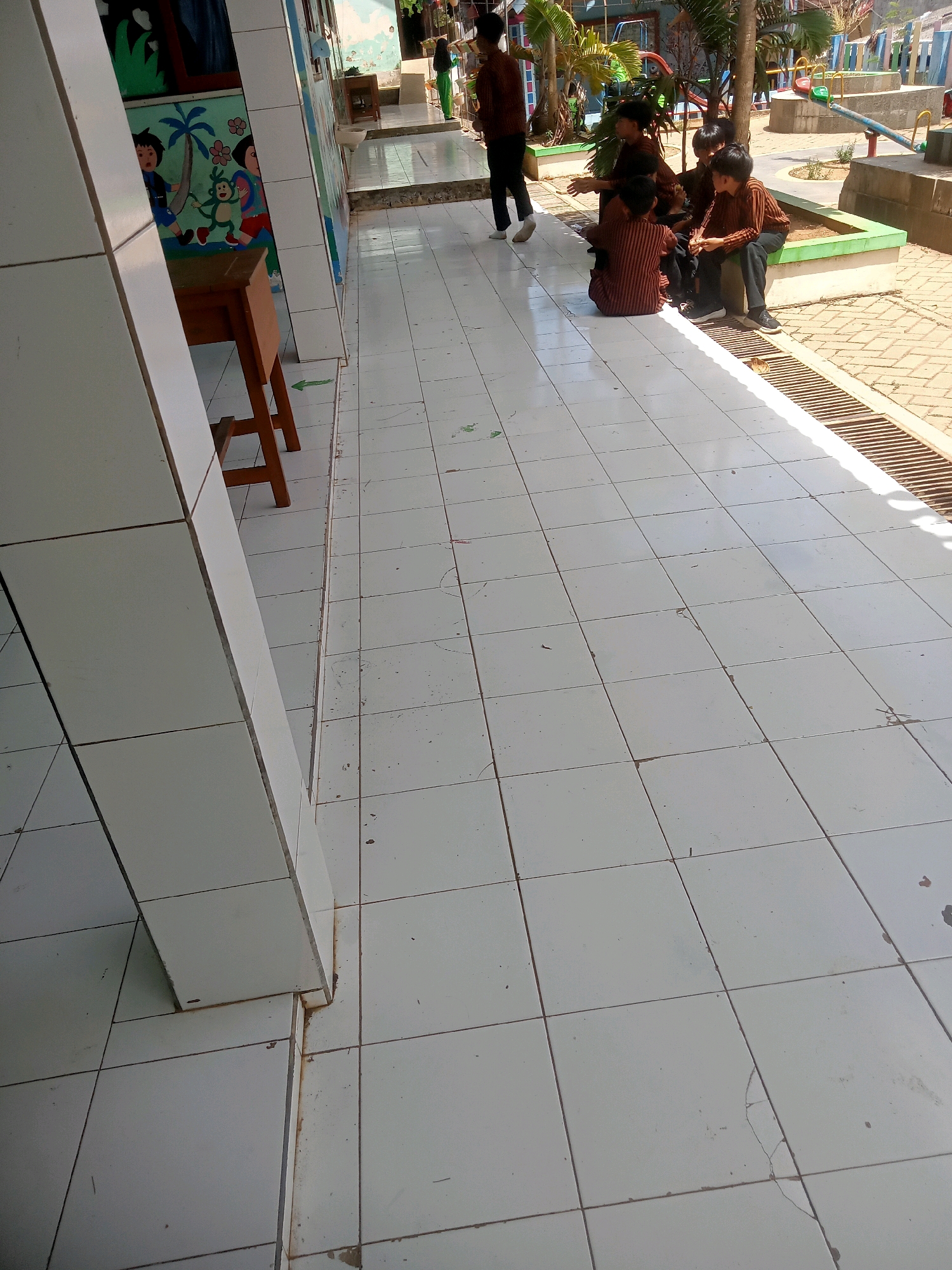 The image depicts a group of young boys, likely students, gathered outside on a tiled walkway. They are all dressed in matching brown and striped uniforms, suggesting they are from the same school or institution. The boys are sitting and interacting with each other, some appearing to be engaged in conversation. The environment appears to be an outdoor area of a school or educational facility, with murals on the wall to the left and playground equipment visible in the background. The location is identified as Pule, Indonesia. The walkway is tiled with white squares and a drainage grate runs along the edge. Sunlight is casting shadows, indicating it is daytime.