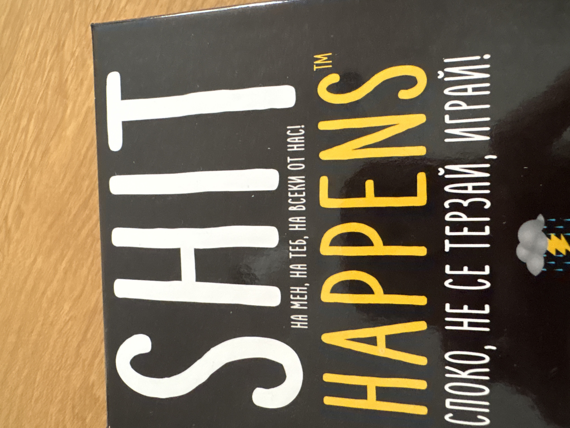 The image shows a close-up of a board game box titled "SHIT HAPPENS". The words "SHIT" and "HAPPENS" are written in large, bold letters, with "SHIT" in white and "HAPPENS" in yellow. There is also text in Bulgarian: "НА МЕН, НА ТЕБ, НА ВСЕКИ ОТ НАС!", which translates to "On me, on you, on everyone of us!". Another Bulgarian phrase "СПОКО, НЕ СЕ ТЕРЗАЙ, ИГРАЙ!" translates to "Calm, don't worry, play!". A small image of a cloud with lightning and rain is also visible. The photo appears to be taken on a wooden surface. The game is likely located in Sofia, Bulgaria, given the presence of Bulgarian text.