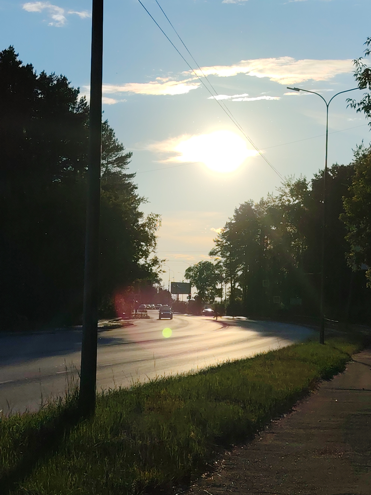 The image depicts a street scene with a road curving to the right, bordered by trees on both sides and a grassy verge. The sun is low in the sky, casting a bright glare that obscures details of the road and creates lens flare. Several cars are visible on the road, driving away from the viewer. A lamppost stands on the right side of the road. Power lines are visible in the sky. The overall impression is of a sunny day, possibly late afternoon or early morning. The location is identified as Chaikovsky, Russia.