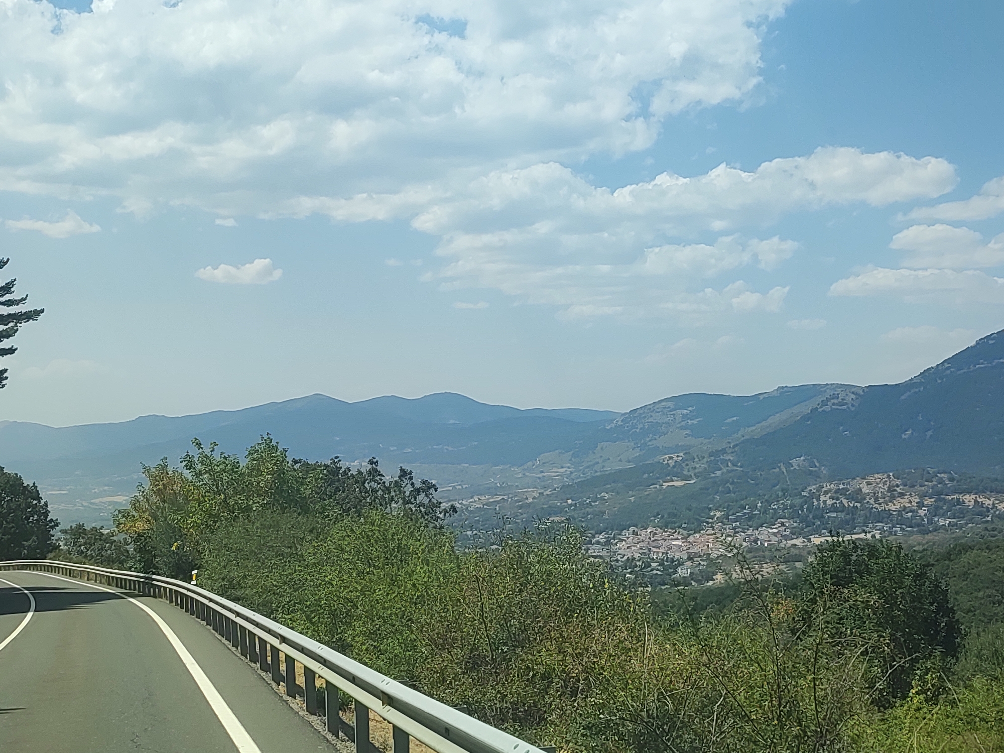 The image captures a scenic view from a winding road, likely in or near Navacerrada, Spain. The road, paved with asphalt, features a white line marking its edge and a silver metal guardrail on the right side. Lush green trees and foliage line the roadside. In the distance, rolling hills covered in vegetation form a layered landscape, with a town nestled in the valley below. The town appears to have buildings with red-tiled roofs. The sky above is a clear blue, dotted with scattered white clouds. The overall scene suggests a mountainous or hilly region, typical of the Sierra de Guadarrama.