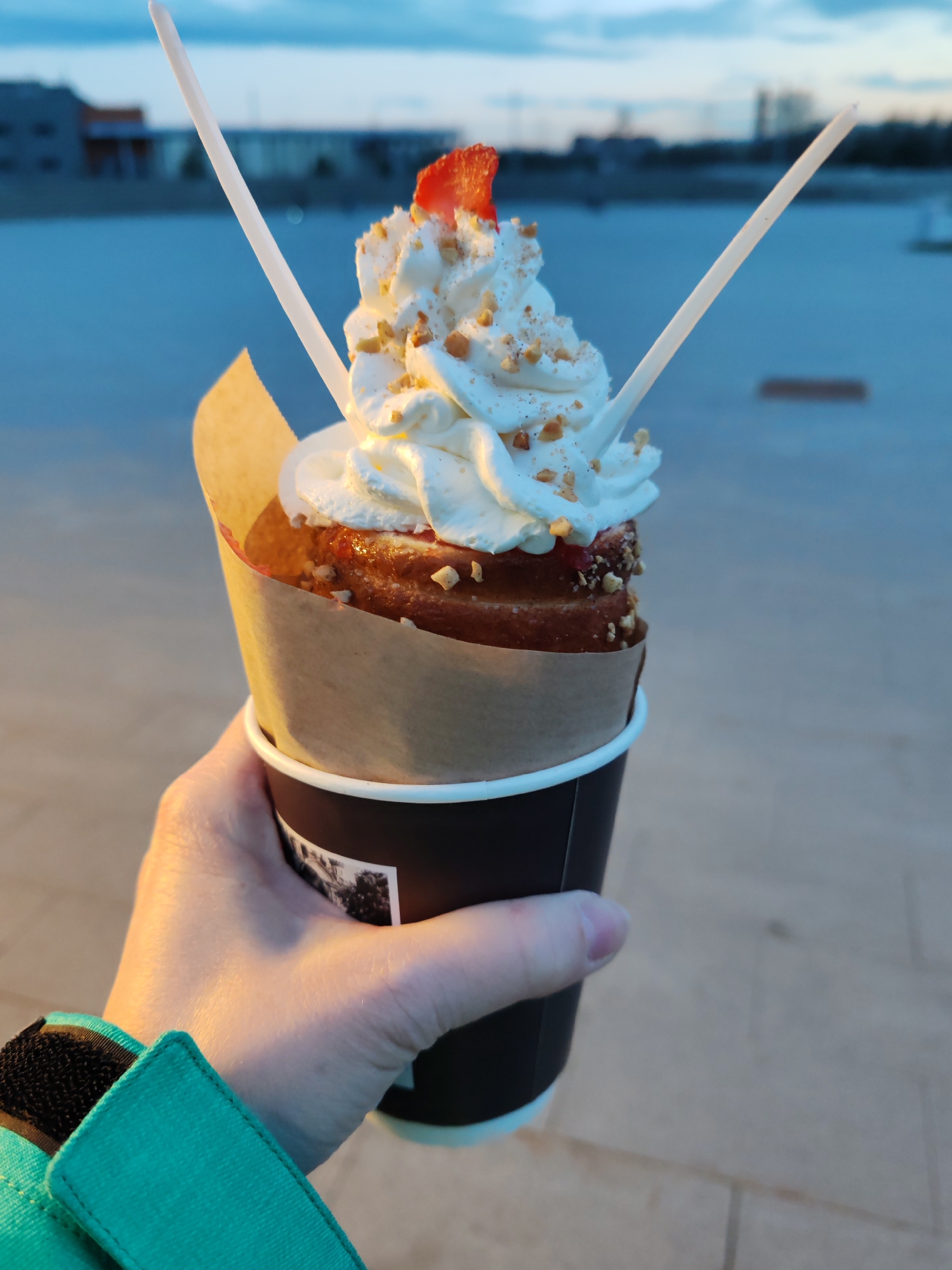 A close-up shot captures a hand holding a dessert outdoors during what appears to be golden hour or dusk. The dessert is a chimney cake (Trdelník) served in a black paper cup with a brown paper sleeve. The cake is filled with a reddish jam or fruit filling and generously topped with swirls of white whipped cream, sprinkled with chopped nuts, and garnished with a small slice of red strawberry. Two slender white stirrers or spoons are inserted vertically into the whipped cream.
The hand, with fingers lightly gripping the cup, wears a teal-colored jacket sleeve with a black Velcro closure. The background is softly blurred, revealing a calm, light blue sky with faint cloud cover and the silhouettes of modern buildings across a body of water, likely a river or canal in Saint Petersburg, Russia. The low sun angle casts a warm, golden glow on the dessert and the hand, creating a serene and inviting atmosphere. The black paper cup features an indistinct, dark grey graphic design on its side, which is not clearly readable. The overall scene suggests a pleasant moment of enjoying a sweet treat by the water.