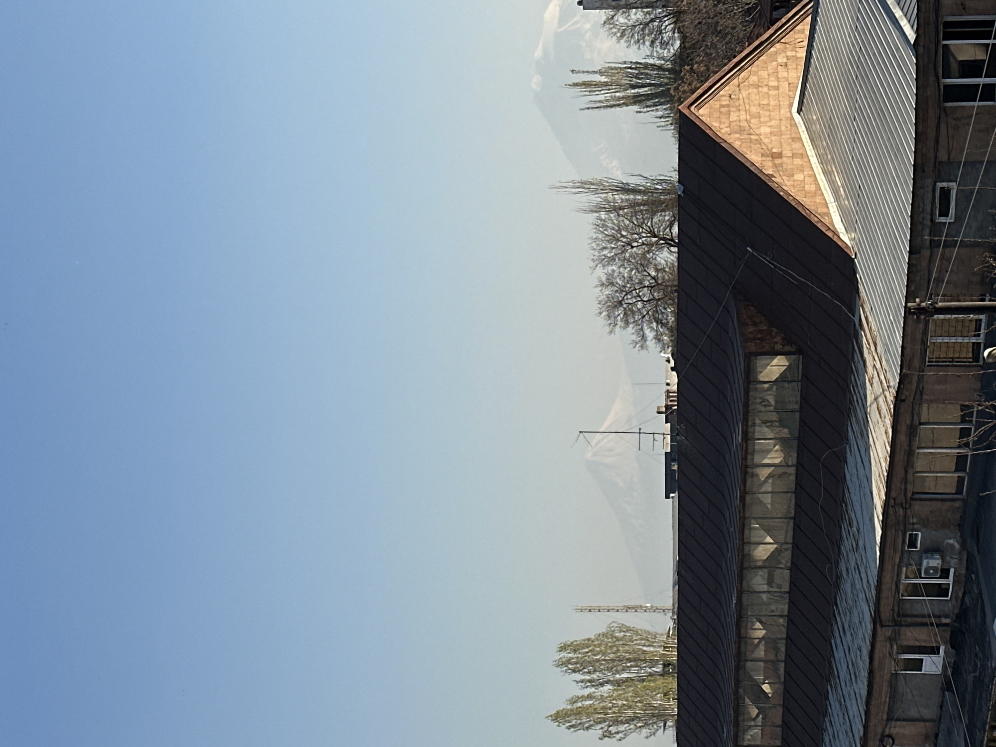 The image captures an urban landscape view under a clear, hazy blue sky. On the right side, a prominent building features a multi-faceted roof and facade, primarily covered in dark brown corrugated metal, with sections of lighter, possibly tiled, roofing. Reflective window strips are visible on one of its walls. Adjacent to it on the far right, an older, somewhat weathered building with a textured facade, several rectangular windows, and an external air conditioning unit is present.
Scattered throughout the mid-ground and background are numerous trees, some with bare branches suggesting late winter or early spring, and others displaying light green foliage. The upper and left portions of the image are dominated by a vast expanse of clear blue sky, which gradually lightens and becomes hazier towards the distant horizon. Faint outlines of mountains or large hills are discernible in the far background, partially obscured by this atmospheric haze. A slender structure resembling a communications mast or antenna can be seen in the distance, barely visible above the treeline.
The scene depicts daytime with strong sunlight, creating distinct shadows on the buildings. There is no specific activity or event taking place; it's a static depiction of a cityscape or urban fringe area with natural elements. No visible text is present in the image.