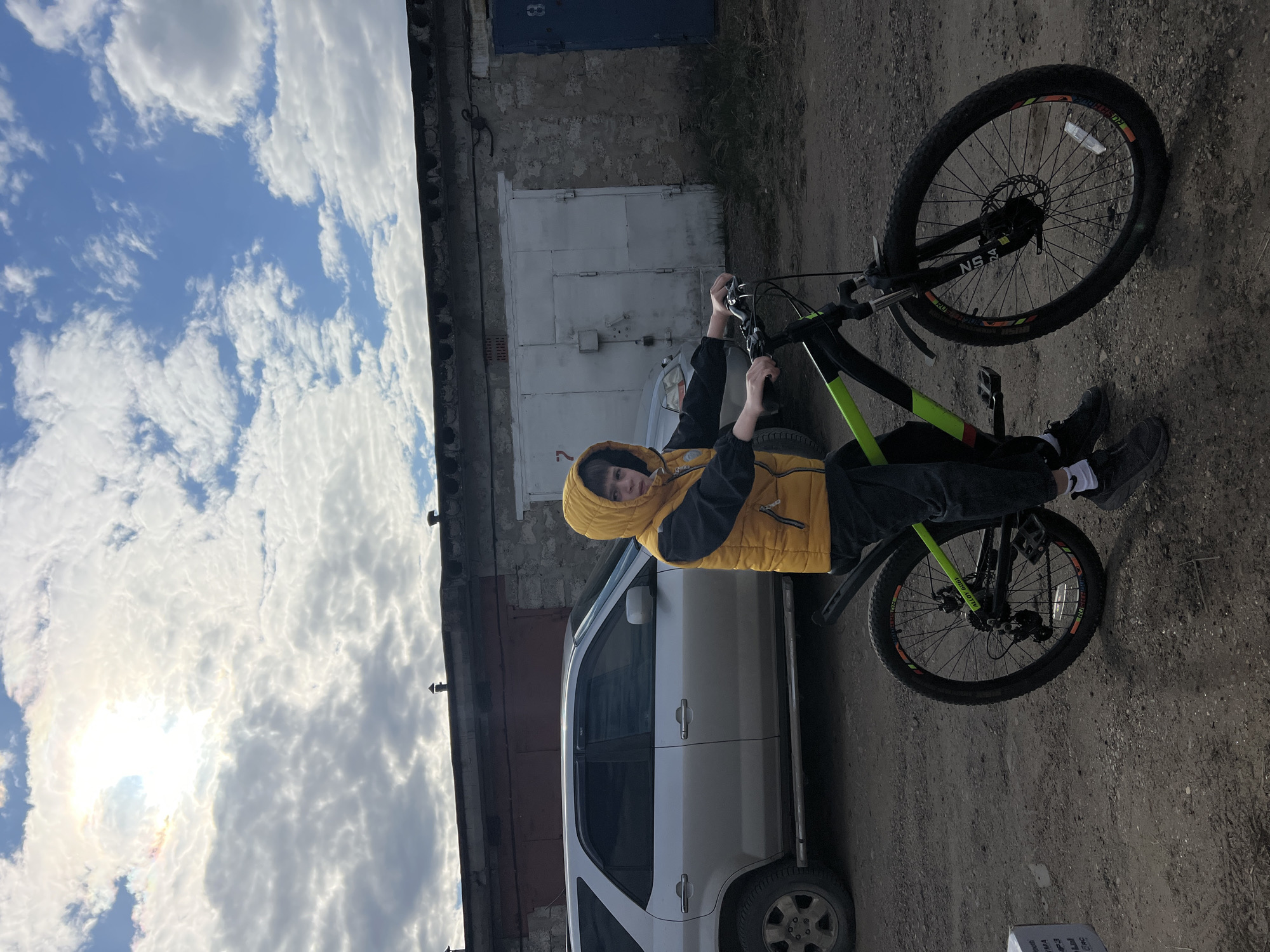 A young person wearing a yellow hooded vest is sitting on a green and black bicycle outside a garage. A silver SUV is parked nearby. The location appears to be Noviy, Russia, based on textual context, although this cannot be confirmed from the image itself. The environment is characterized by overcast skies and a dirt ground.