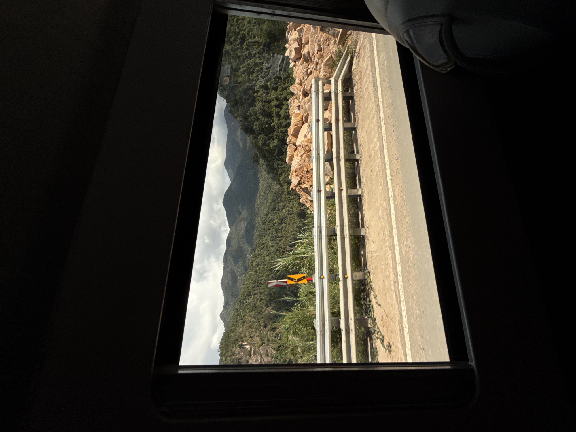This media file captures a scenic view from a window, likely of a vehicle, looking out onto a mountainous landscape. The frame of the window is dark, contrasting with the brightly lit exterior. The scene is oriented vertically in the image, suggesting it was taken with the camera rotated.
The main subjects are the natural environment and man-made structures along a road. A winding road is visible, bordered by a metal guardrail. Beyond the road, a steep, rocky slope covered in dense green trees dominates the left side of the frame. In the distance, rolling mountains fade into a cloudy sky. A traffic sign, yellow with a black arrow indicating a sharp turn, is prominently placed near the roadside vegetation.
The time of day appears to be late afternoon, with long shadows cast by the sun hitting the mountains and road. The weather seems partly cloudy, with sunlight breaking through. The location, Nam Khanh Vinh, Vietnam, is hinted at by the lush, tropical vegetation and mountainous terrain. There are no people visible in the frame, and the overall mood is one of natural beauty and peaceful travel. The only visible text is on the traffic sign, which is a directional arrow.