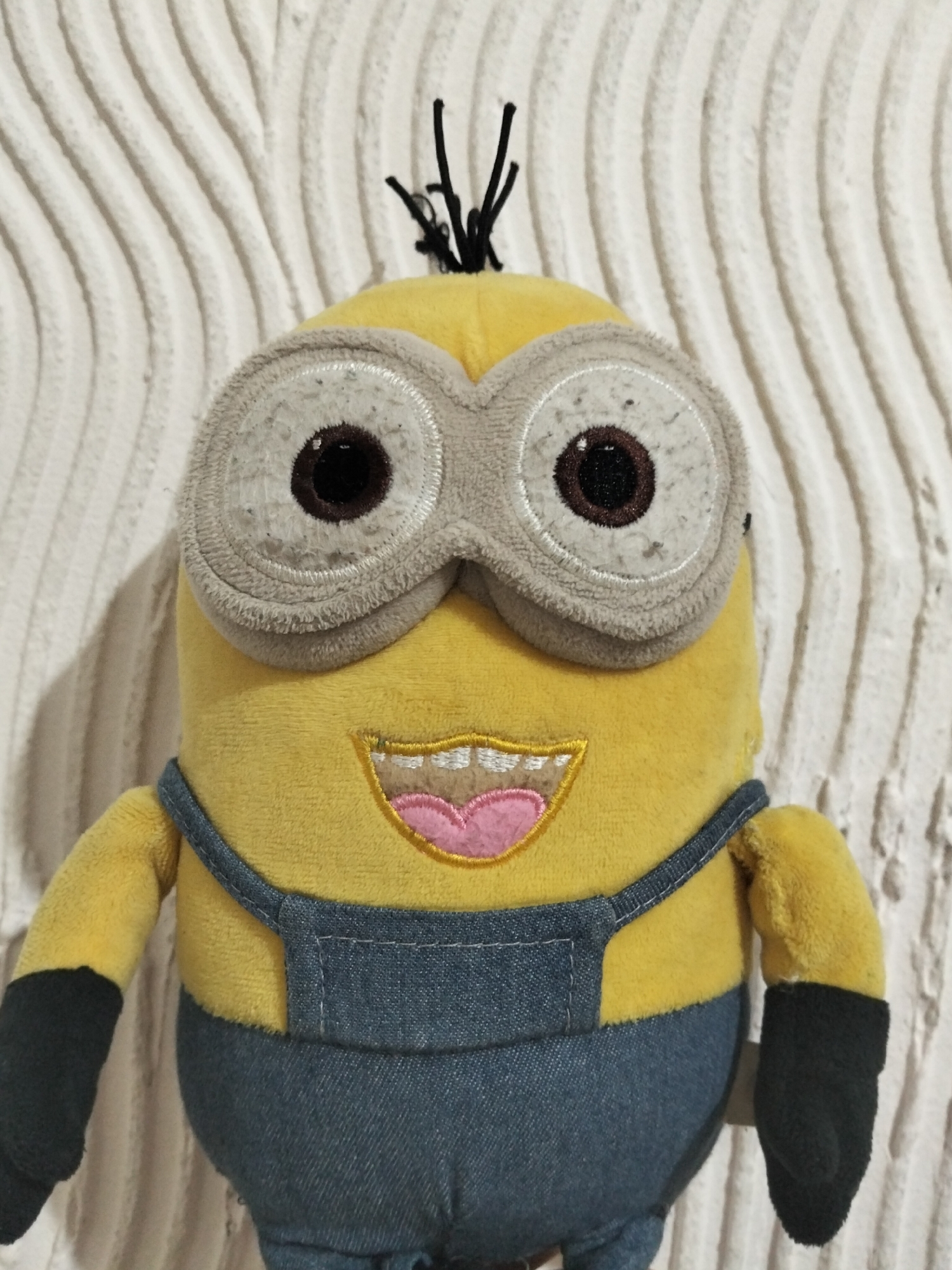 A plush toy minion, with a yellow body, blue denim overalls, and large goggles, is positioned in front of a white, textured wall. The minion has one eye, a wide smile with visible teeth and a pink tongue, and a few strands of black hair sticking out from the top of its head. Its arms are black. The background wall features a pattern of parallel, wavy ridges. The lighting is diffuse, casting soft shadows. The location is Unknown, Unknown.