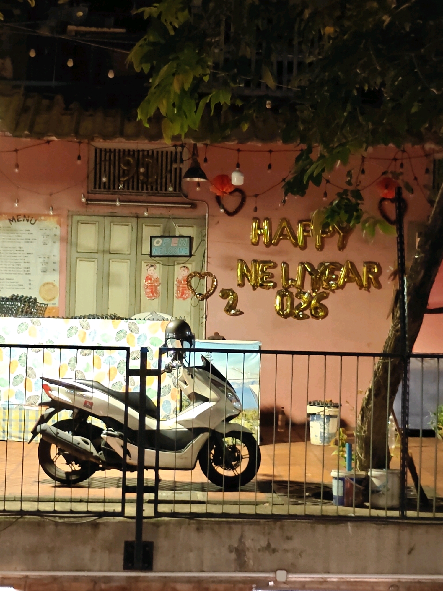 The image captures a storefront in Bangkok, Thailand, at night. A pink-colored building is visible, adorned with gold foil balloons that spell out "HAPPY NEW YEAR 2026". String lights with small bulbs and heart-shaped ornaments hang across the facade. To the left, a "MENU" sign is posted on the wall above a counter. A light green double door is centrally located, with an "OPEN WELCOME" sign above it. Two decorative figures resembling children in red outfits are affixed to the wall near the door. In front of the counter, a white scooter with a black helmet is parked behind a black metal fence. The scooter is partially obscured by the fence. The ground in front of the scooter appears to be paved, with a concrete curb in the foreground. A tree trunk is visible to the right of the scooter. Two buckets, one white and blue, are placed on the ground behind the fence.