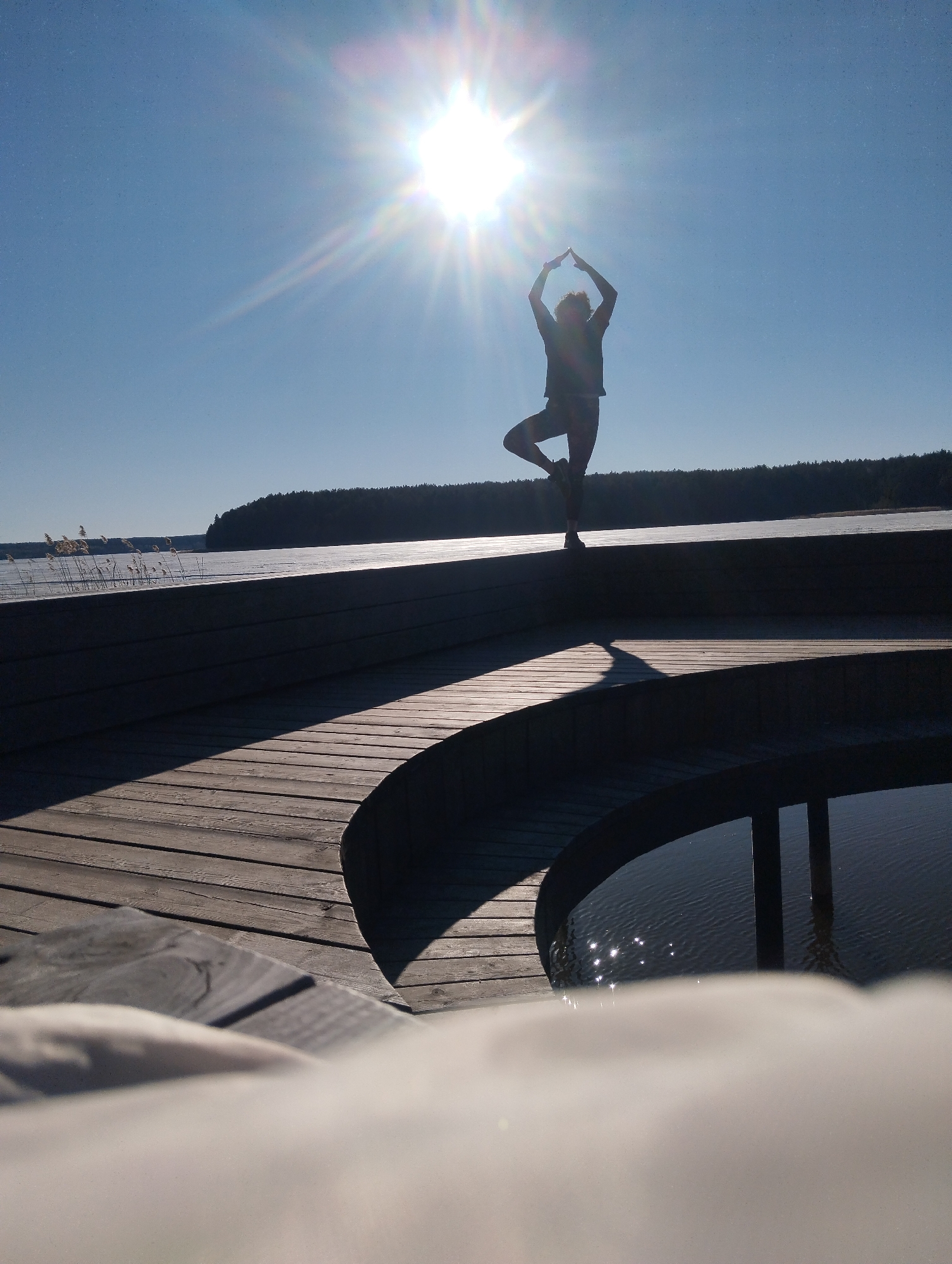 A silhouette of a person is performing a yoga pose on a wooden platform outdoors. The person is standing on one leg with their other leg bent and placed on the inner thigh of the standing leg. Their arms are raised above their head, with their hands joined. The sun is positioned directly above the person, creating a bright glare and causing the person to appear as a dark silhouette. The platform they are standing on has multiple levels and curved edges. In the background, there is a body of water that appears to be partially frozen. Behind the water, a dense forest stretches along the horizon. The sky is clear and blue. The location is Unknown, Unknown.