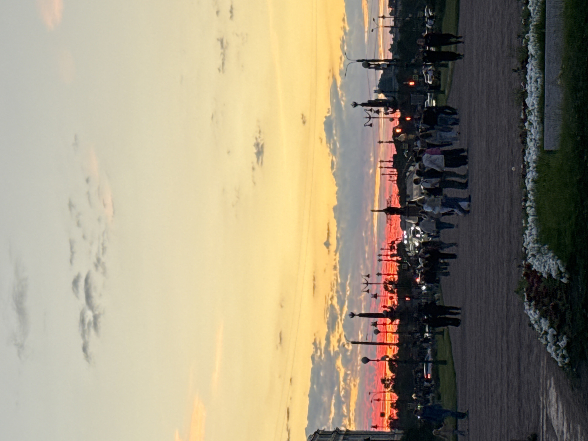 The image depicts a sunset over the city of Saint Petersburg, Russia, likely viewed from a park or public space. The sky is filled with vibrant hues of orange and yellow, with a band of intense red just above the horizon, indicating the setting sun. Wispy clouds are scattered across the sky, tinged with pink and gray.
In the foreground, a wide, paved walkway or plaza is visible, with a low-lying flowerbed filled with white and green foliage in the bottom right corner. A group of people are walking along the pathway, silhouetted against the bright sky. They are dressed in casual clothing, and their poses suggest they are strolling or gathering. To the left, a person with a dog is also walking along the path.
Beyond the walkway, the cityscape is visible. The silhouettes of buildings, streetlights, and trees form a dark line against the colorful sky. Several ornate streetlights with multiple lamps are prominent. In the distance, the outlines of some taller structures, possibly monuments or significant buildings, can be discerned. Cars are also present on a road in the mid-ground. The overall atmosphere is serene, with the warm colors of the sunset creating a peaceful ambiance.