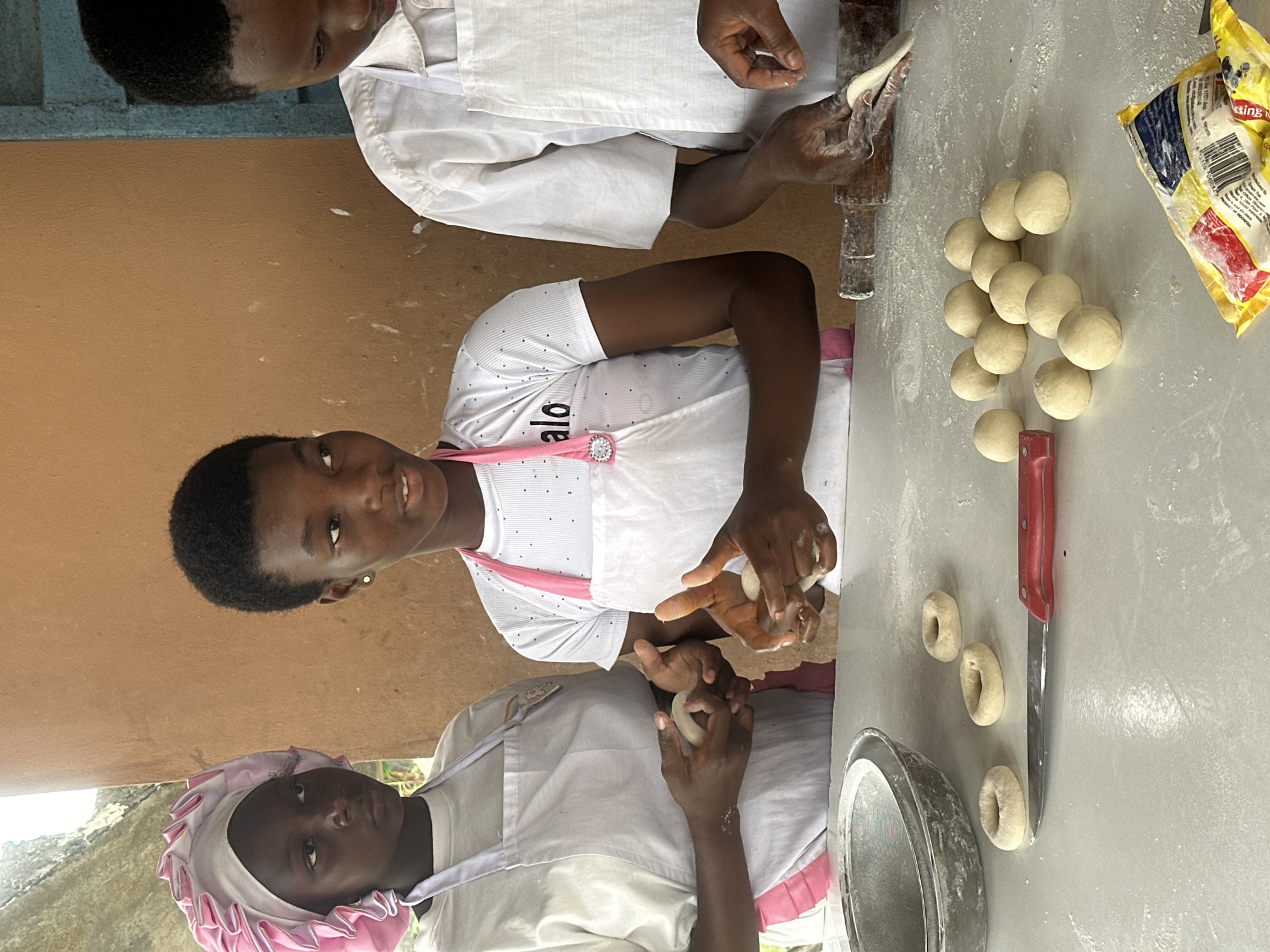 Three young people, likely teenagers, are engaged in preparing dough for baking. The setting appears to be an open-air or poorly enclosed space with a rough, brown wall as the backdrop.
On the left, a person wearing a white apron and a pink and white ruffled head covering is holding a small ball of dough in their hands, seemingly shaping it. To their right, another person, also in a white apron with a pink strap, is actively working with a ball of dough, their hands demonstrating a shaping action. The third individual, on the far right, is wearing a white apron and is partially visible, with their hands in the process of rolling or shaping dough with a wooden rolling pin.
The surface in front of them is a white table dusted with flour. Several small, ring-shaped pieces of dough, possibly early stages of making doughnuts or similar baked goods, are arranged on the table. A red-handled knife lies next to these rings. Further to the right, a collection of uniformly sized, round dough balls is neatly arranged. A yellow packet, possibly containing baking ingredients, is also visible in the lower right corner.
The overall impression is one of a practical, hands-on lesson or activity involving food preparation, possibly in a community setting or an educational program in Badagry, Nigeria.