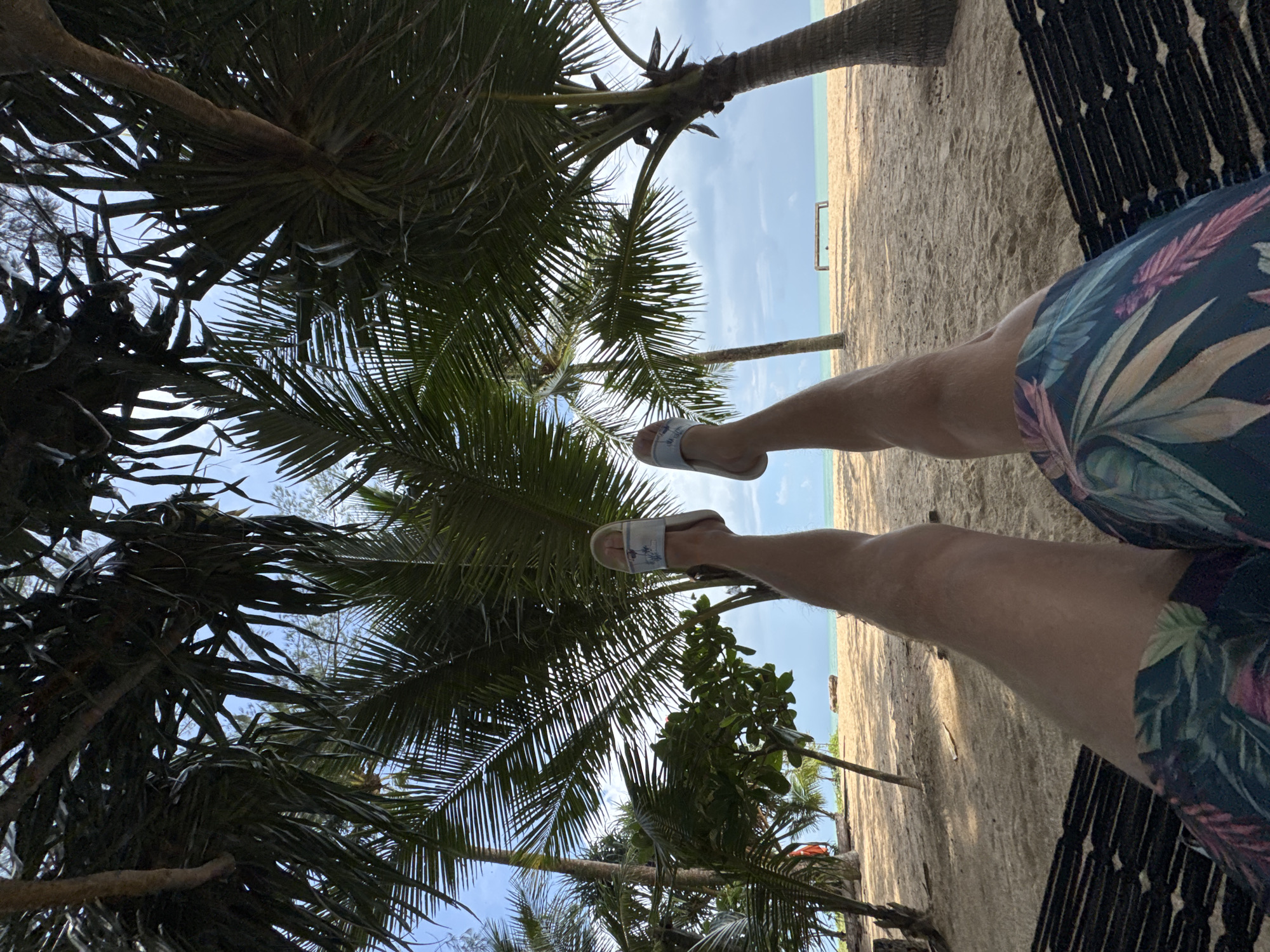 A first-person perspective shot shows a person relaxing in a hammock on a sandy beach, likely in Ban Khuk Khak, Thailand. The person's legs are extended, wearing floral-patterned shorts and light-colored flip-flops. Palm trees with lush green fronds frame the scene from above, casting shadows on the sand. In the background, the turquoise ocean meets a cloudy sky. The beach appears uncrowded, with a wooden structure or bench visible in the distance near the water. The overall atmosphere is one of tropical tranquility.