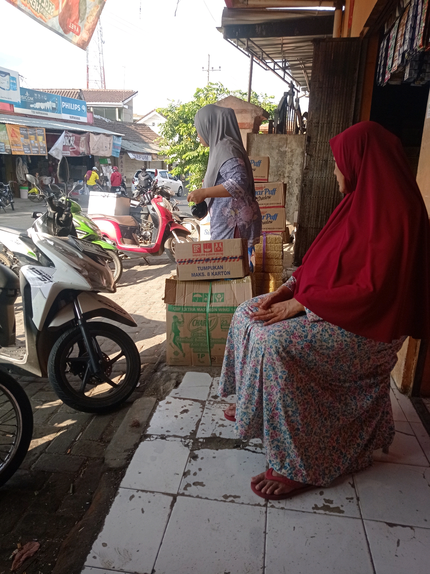 A side view shows two women in hijabs near shops in Tajinan, Indonesia. A woman on the right, wearing a red hijab and a floral dress, sits on a low stool or crate, facing left. Her hands are clasped in her lap, and she wears red flip-flops. To her left, a woman wearing a grey hijab and a patterned top stands, holding a dark bag. In front of the standing woman are several stacked cardboard boxes, some with text visible, including "Charl's." Behind the boxes, a bright red scooter and a white scooter are parked on a tiled walkway. To the left of the scooters, a white motorcycle is parked. In the background, a busy street is visible with more shops, including ones with signs for "Philips" and "ISICOM," as well as more parked motorcycles and a white car. Trees and buildings frame the scene under a bright sky.