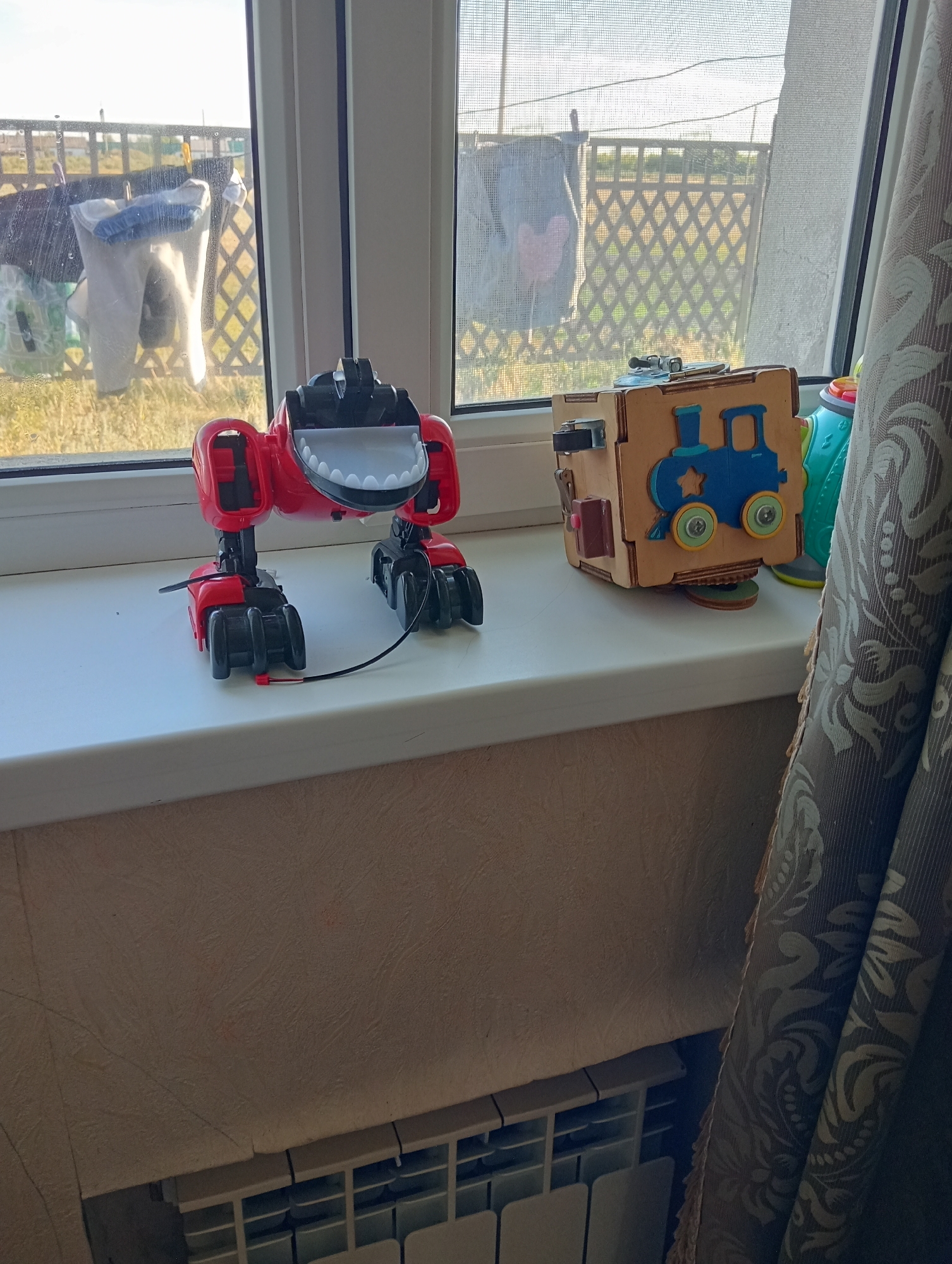 On a windowsill, a red toy dinosaur with black accents stands to the left, facing the viewer. To the right of the dinosaur is a wooden sensory cube with various interactive elements, including a blue train graphic. The scene is captured indoors, with a window in the background showing a wire fence, some laundry hanging on a clothesline, and vegetation. A patterned curtain hangs on the right side of the image. Below the windowsill, a white radiator with multiple horizontal slats is visible. The overall setting appears to be residential.
