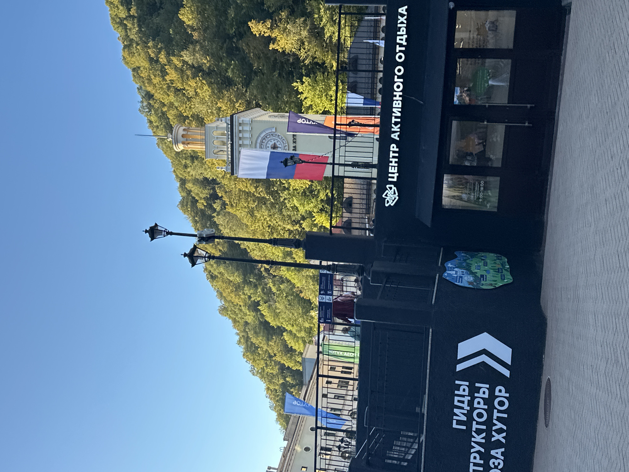 The image shows a street scene in Estosadok, Russia, with a clear blue sky and a forested hillside in the background. In the foreground, a dark, modern building with large glass windows has the text "ЦЕНТР АКТИВНОГО ОТДЫХА" (Center for Active Recreation) written in white across its facade. To the left of this building, a dark wall features white lettering including "ГИДЫ" (guides) and "СТРУКТОРЫ" (instructors). A large white arrow points to the right.
Above the "Center for Active Recreation" building, a tall, light-colored tower with arched windows and a spire stands prominently. The Russian flag is draped over the side of this tower. Several lampposts, characteristic of older European streetlights, are visible along a railing. Behind the railing, there are buildings with windows and balconies, and people can be seen walking. In the distance, the hillside is densely covered with green and yellow foliage, indicating autumn. The ground in front of the building appears to be paved with light-colored stones.
