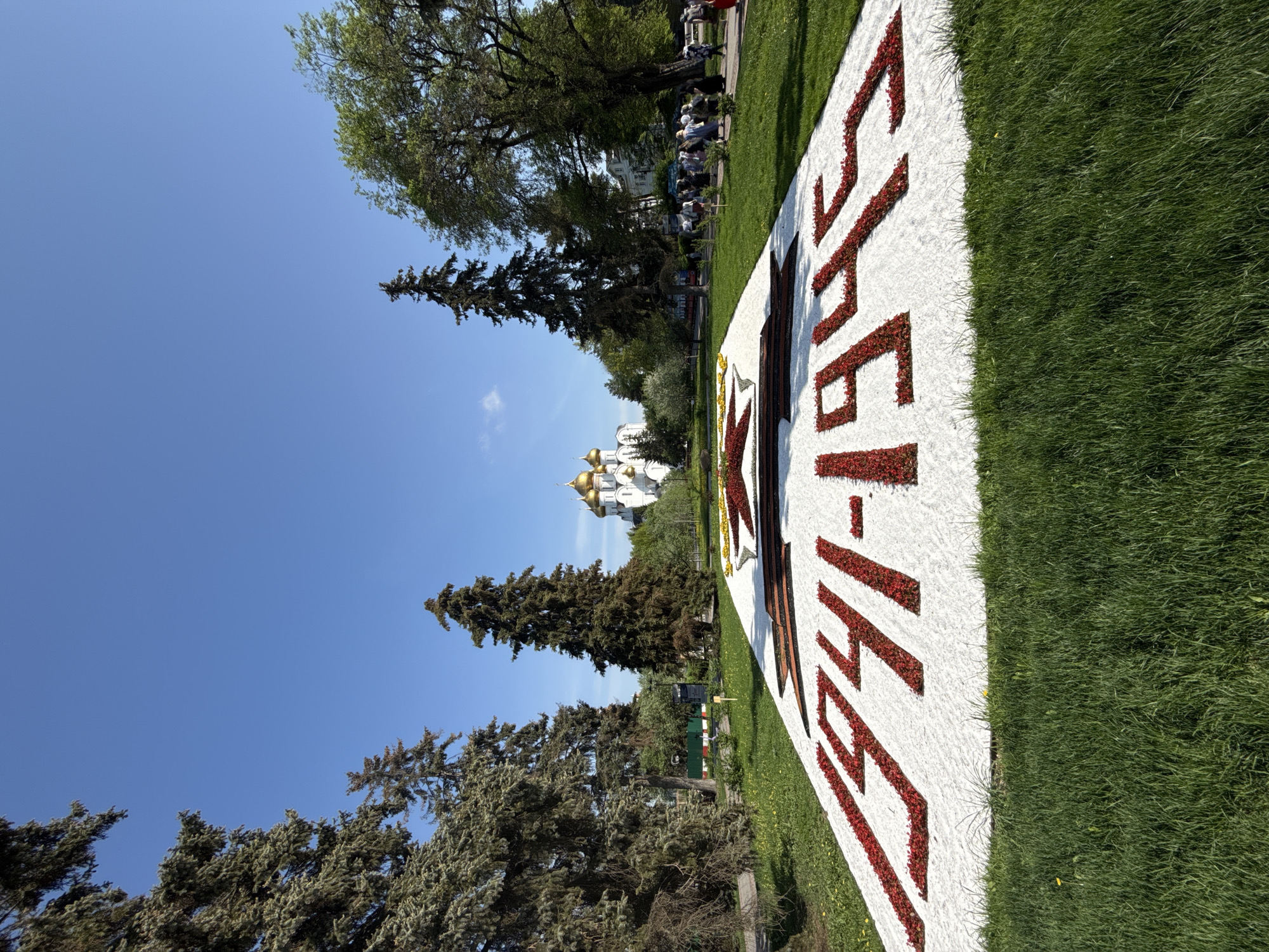 A slightly low-angle, medium-distance shot features a large flower display in the foreground with a church in the background, set against a bright blue sky.
The flower display is predominantly white with red accents, displaying the date "1941-1945". This display is set in a grassy area.
In the background, a white-walled church with golden domes is visible. The building architecture is notable, especially the multiple onion-shaped domes characteristic of Russian Orthodox churches.
A cluster of people is visible in the background, near trees on the edge of the lawn.
The environment is mostly sunny, with trees providing a backdrop to the scene. The location appears to be a public space in a city, although the specific city is unknown.