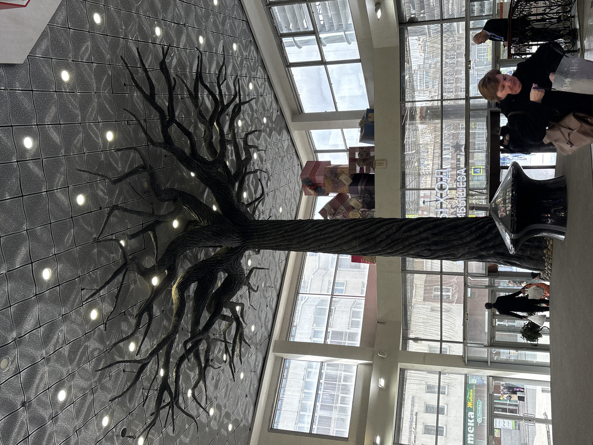 The image captures an interior view of a shopping mall in Yekaterinburg, Russia, featuring a decorative tree sculpture extending from the wall to the ceiling. The wall is textured with a grid pattern and illuminated by embedded lights. The mall's interior is visible through large windows, revealing shops and pedestrians. Two people are near the windows, one is looking toward the interior. Another individual is walking into or out of the building. Other shopping materials and signage are apparent in the background. The environment suggests a commercial space during the day.
