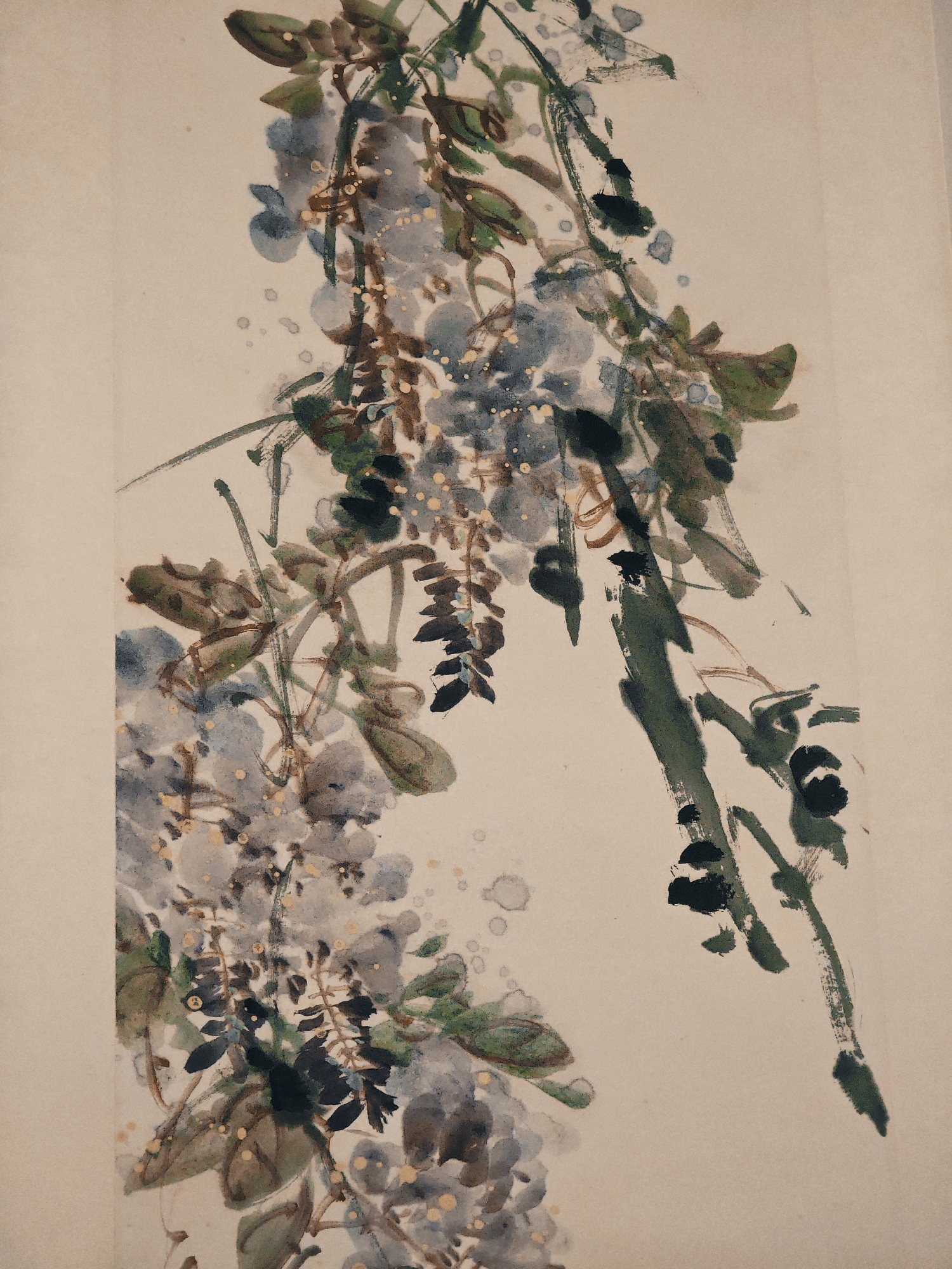 The image displays a vertical close-up of a traditional East Asian ink wash painting, depicting flowering wisteria branches and foliage. The artwork is characterized by dynamic brushstrokes and varied washes of color against a plain, light beige background.
The primary subjects are long, cascading clusters of wisteria flowers rendered in soft blue-grey tones, interspersed with various shades of green foliage and brown-black branches. The leaves are depicted with expressive, fluid strokes, showing different levels of detail and ink saturation. Small, light yellow or golden splatters and dots are scattered across the painting, particularly among the flower clusters, which could represent pollen, dew, or a decorative embellishment, adding a subtle shimmer.
The composition highlights the natural elegance of the plant, with branches and flower clusters extending diagonally across the frame, creating a sense of organic movement. The background is simple and unadorned, emphasizing the botanical subject. No human figures, specific scenes, or discernible activities are depicted. The artistic style suggests an East Asian origin, likely Chinese, given the provided location context. The texture of the paper or silk medium is subtly visible, reflecting the traditional craftsmanship. No visible text is present within the artwork.