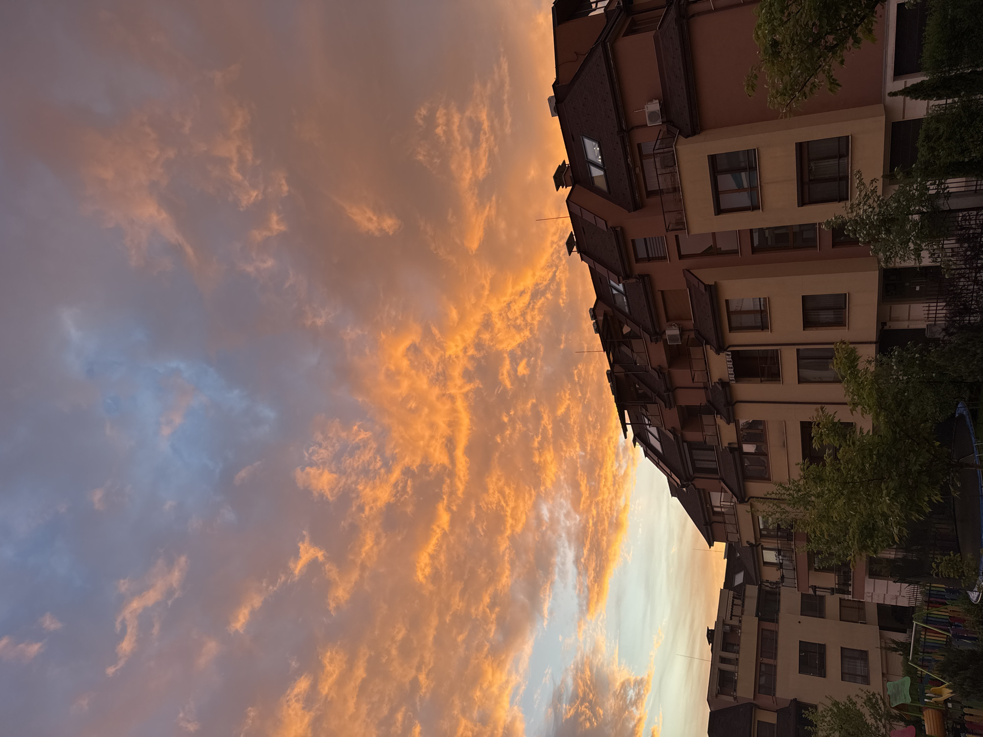 A vertical, long shot depicts a building alongside an open area in what appears to be a city. The architecture is modern, with multi-story buildings having various windows, balconies, and rooftops. Trees are interspersed between the buildings. A complex sky of clouds stretches across the top of the image, ranging from shades of orange to pale blue. The location could be in Sofia, Bulgaria.