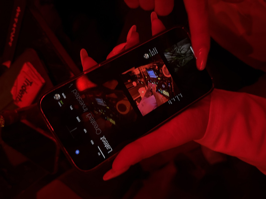 The image captures a close-up view of a person's hands holding a smartphone in a dimly lit environment, saturated with red light. The setting appears to be indoors, possibly a club or bar, given the dramatic lighting and visible background elements.
The hands, primarily visible in the center-right of the frame, have manicured nails painted in a light color (appearing reddish due to the strong red light). The fingers are curved around the sides and back of the phone, holding it horizontally in landscape mode.
The smartphone screen is illuminated and displays content from what appears to be a social media or content browsing application set in dark mode. The text on the screen is mostly in white or light gray. Prominently displayed is inverted text that reads "Latest Created Invested 1" with an underline beneath "Latest". Below this, there are columns of numerical values such as "300k" (under "Created") and "500k" (under "Invested"), suggesting counts or metrics related to these categories. On the far left of the screen, smaller text and icons are visible, including a generic profile picture icon, "06:30", "06:20", "New", "Today", and "20/09", possibly indicating timestamps or recent activity.
The main visual content on the screen consists of two visible thumbnails. The larger and more central thumbnail shows an image or video frame of a person (likely a DJ) interacting with professional DJ equipment, including a mixing console with turntables/jog wheels, numerous buttons, and glowing lights, indicating a music-related context. A smaller thumbnail in the upper right corner of the screen displays what looks like green foliage or a plant.
In the blurred background to the left of the phone, a white packet or box of "Trident" gum is partially visible, oriented upside down. Other dark, indistinct objects, possibly bottles or containers, reflect the red light. The overall activity depicted is a person browsing content on their smartphone, likely engaging with a platform that features user-generated content or investment-related feeds, under ambient red lighting.