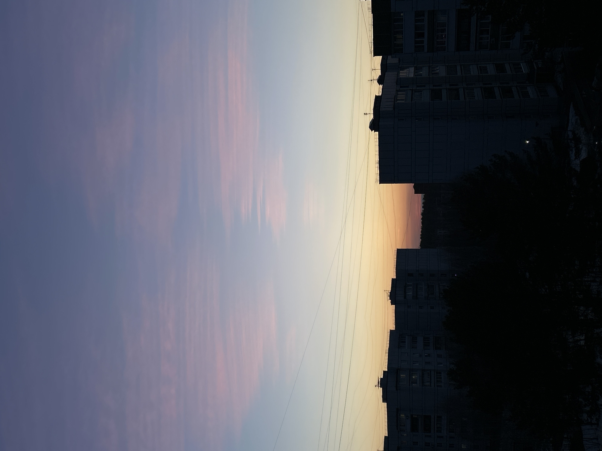 The image depicts a twilight scene in what appears to be a residential area. The sky exhibits a color gradient from purple-pink to light blue and yellow, indicative of either sunrise or sunset. Silhouetted against this sky are apartment buildings, likely located within an urban environment. Overhead power lines are visible, stretching across the skyline. The scene lacks clear indications that it is in Chaikovsky, Russia, beyond the architectural style of the buildings. The overall image is taken in portrait orientation. The base of the frame is dark and obscured by the buildings and potential foliage. No people are visible in the image.