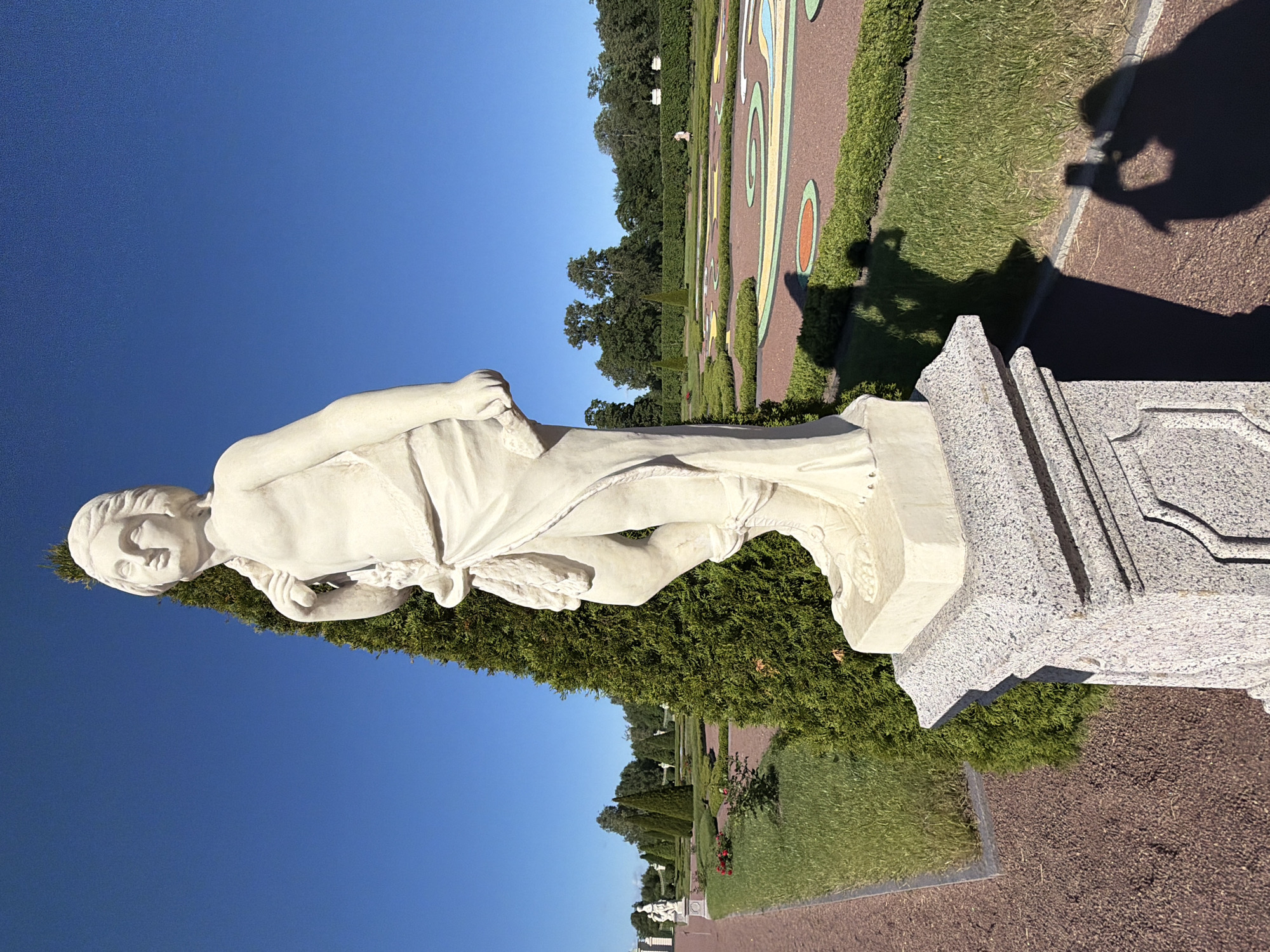 This is a photograph of a stone statue of a person in a garden setting. The statue is light-colored and appears to be made of marble or a similar material. It depicts a person with flowing drapery, possibly a mythological figure or a historical representation. The statue is positioned on a pedestal made of speckled granite.
The background of the image shows a well-maintained garden with manicured hedges, green grass, and pathways. There are colorful patterns on some of the pathways, which suggest a formal garden design. In the distance, there are trees and a clear blue sky. The lighting indicates it is daytime, likely morning or afternoon, with bright sunlight casting shadows. The shadow of a person holding a camera is visible in the lower right portion of the frame, indicating someone is taking the picture.
The overall mood of the image is serene and picturesque, highlighting the beauty of the statue and the garden. There is no visible text in the image. The location is not specified but appears to be an outdoor public space or a private estate with extensive landscaping.
