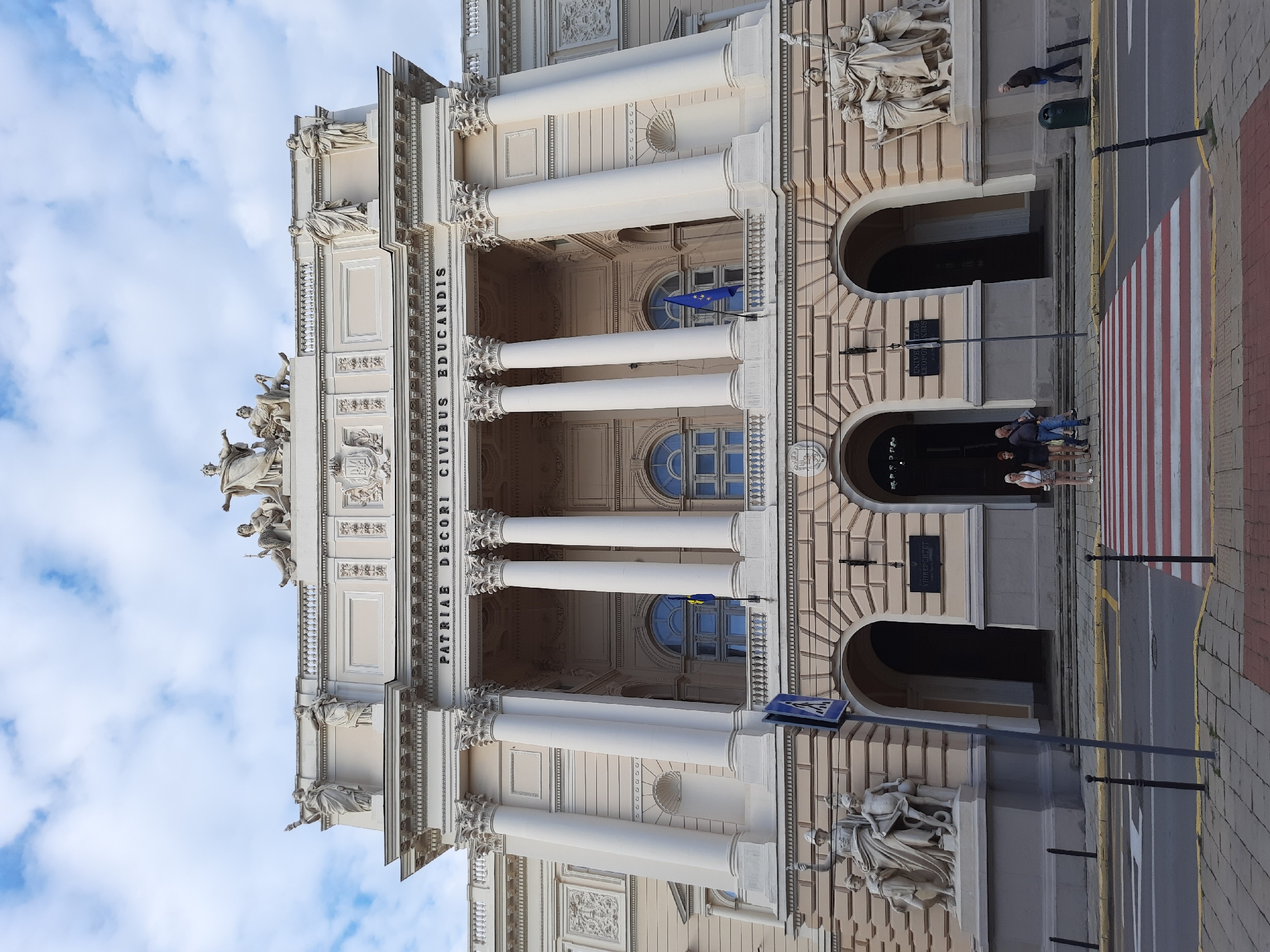 The image shows the front of the Ivan Franko National University of Lviv in Lviv, Ukraine. The building is a large, ornate neoclassical structure with a central portico supported by Corinthian columns. Statues adorn the facade and roofline. In front of the building, there is a pedestrian crossing with red and white stripes. Several people are visible: a group of three individuals standing on the sidewalk near the entrance, and another individual walking across the street. A blue and yellow flag, likely the Ukrainian flag, is visible on a balcony. The sky is partly cloudy.