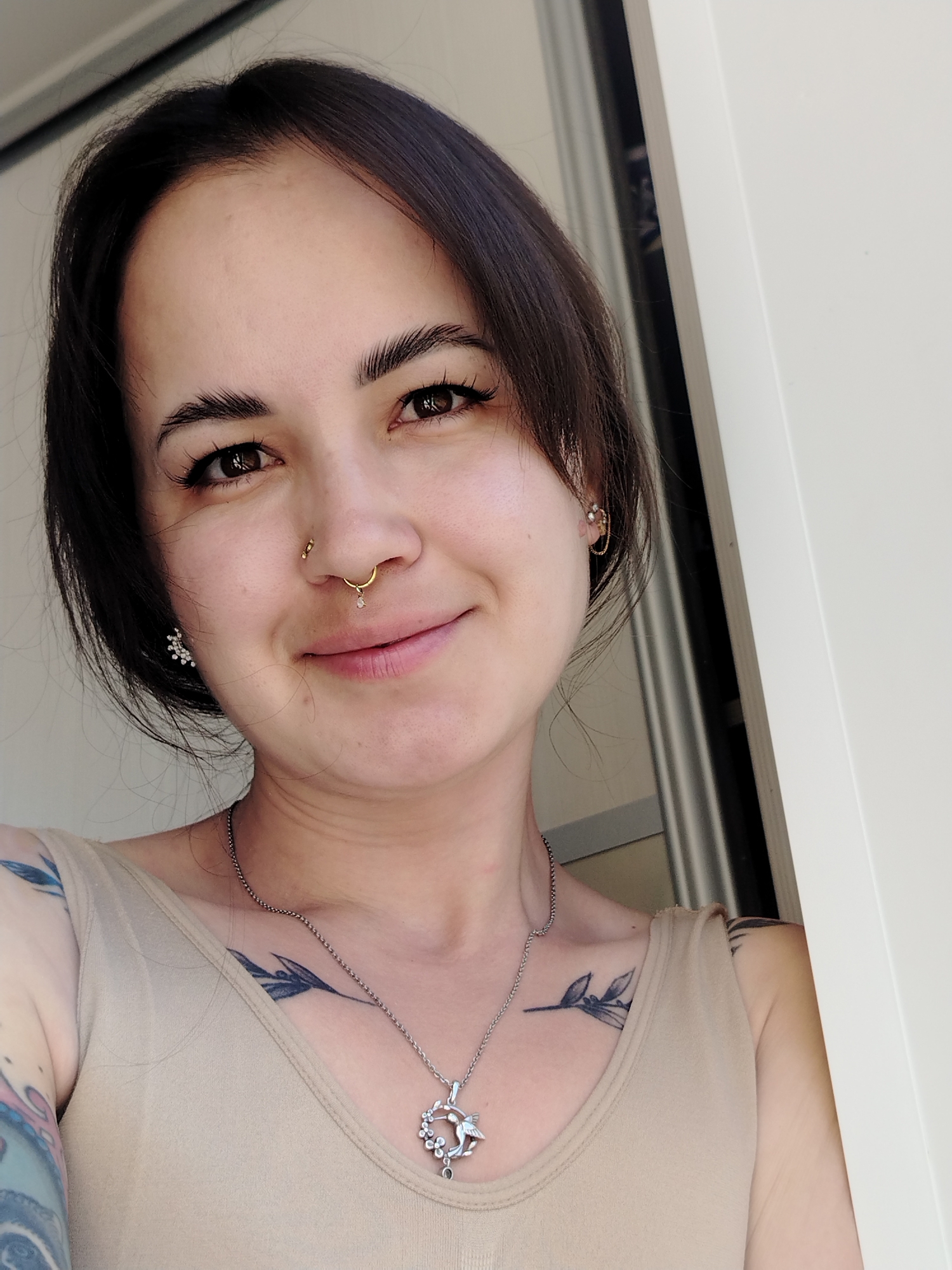 A medium shot depicts a woman with fair skin, dark hair styled around her face, and brown eyes. She is smiling. She has a nose ring, earrings, and a necklace with a pendant. Tattoos are visible on her shoulders and arm. She is standing near a white doorframe. The location is not discernible as Noviy, Russia.