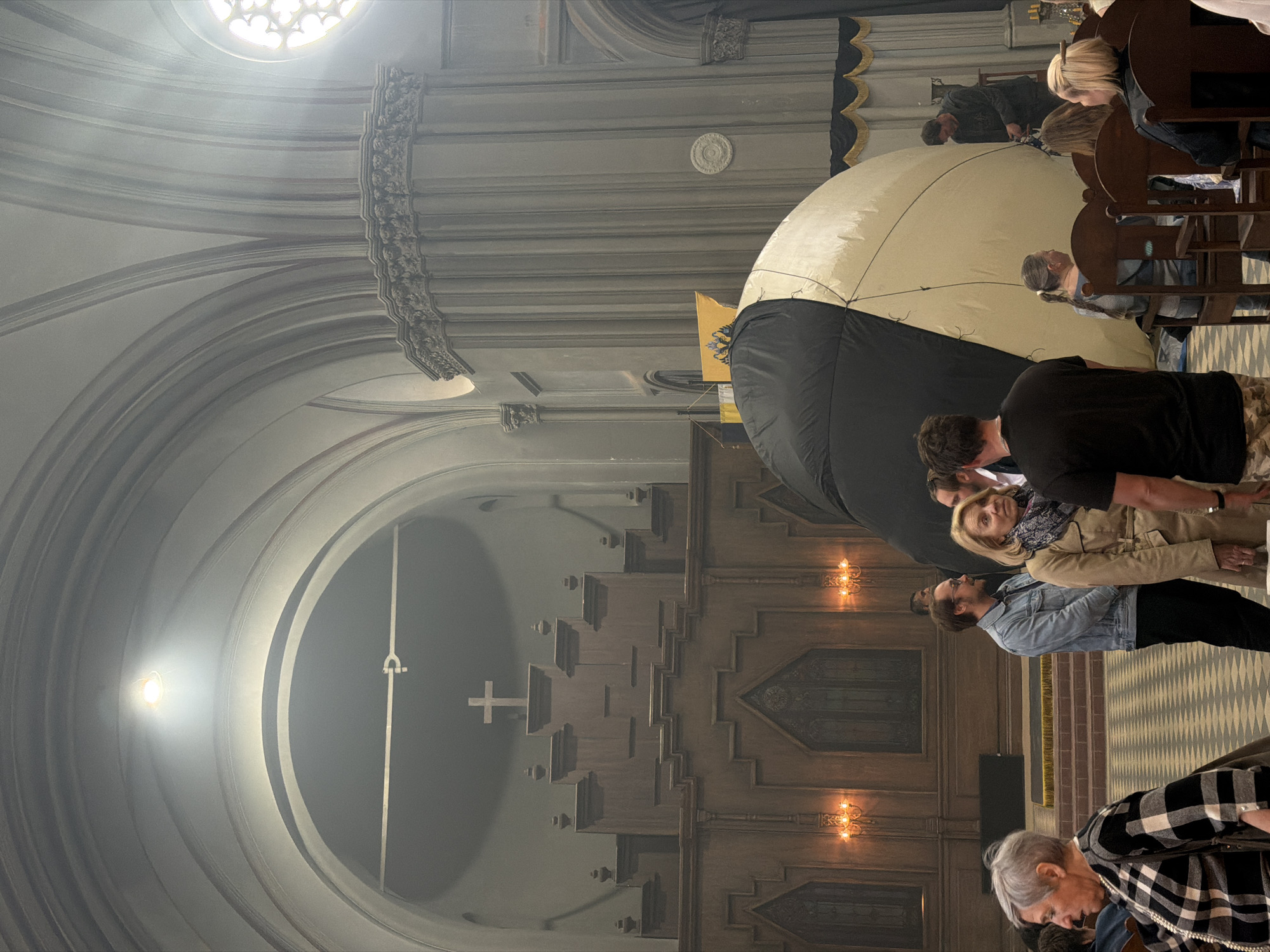 The image captures an indoor scene within a grand, historically rich building, likely a church or cathedral, situated in Saint Petersburg, Russia. The setting is characterized by its impressive architecture, featuring high vaulted ceilings, intricate arches, and ornate decorative moldings in shades of grey stone. A large, circular rose window, high on the left, serves as a primary light source, casting bright light into the space. Further into the background, a simple white cross is mounted on a wall. The right side of the image showcases rich, dark wooden paneling with detailed carvings and two illuminated wall sconces, suggesting a blend of gothic and classical design elements.
A central element of the scene is a large, spherical or semi-spherical inflatable object, divided into a black lower half and a light-colored (beige/white) upper half. This object appears to be an art installation or a temporary exhibition piece within the sacred space.
Several individuals are present, observing the surroundings. Some people are standing in the foreground and mid-ground, dressed in casual attire such as a denim jacket, plaid shirt, and t-shirts, looking towards the inflatable object or generally taking in the environment. Others are visible sitting in traditional wooden pews on the far right. The overall activity suggests attendees at a public viewing or a special event. The time of day appears to be daytime, with natural light entering from the high window, contributing to a generally dim yet reverent atmosphere. No visible text is discernable in the image.