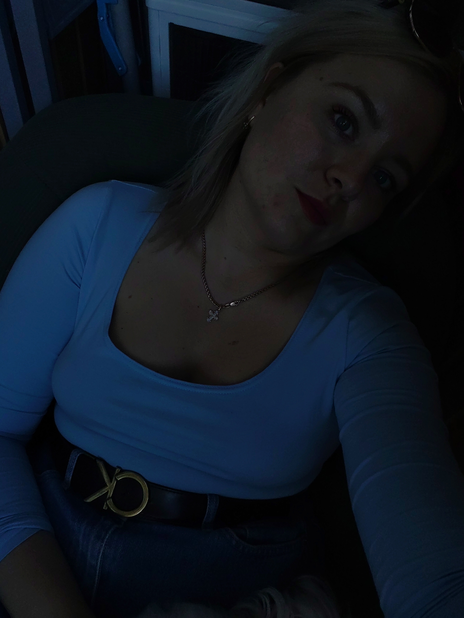 In a dimly lit indoor environment, likely a residence or vehicle, a young Caucasian woman with blonde hair is seated and looking towards the camera. She is wearing a light blue long-sleeved top and blue jeans, cinched with a black belt featuring a gold buckle with the "CK" logo. A silver chain necklace with a small cross pendant adorns her neck. The background is dark and indistinct, with hints of furniture or objects that are not clearly identifiable. The lighting suggests it is nighttime or the interior is poorly illuminated. No specific landmarks of Izhevsk, Russia, are visible.