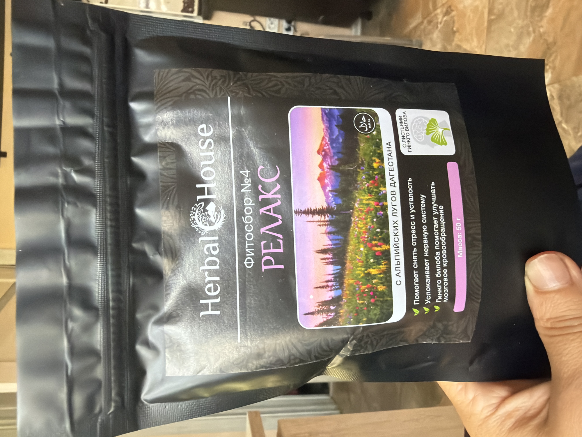 The image shows a black resealable pouch of "Herbal House" brand herbal tea, labeled "Фитосбор №4 РЕЛАКС" (Herbal Blend No. 4 RELAX). The pouch features a scenic image of a mountain landscape with a field of wildflowers at sunset. Text on the pouch indicates that it is "С АЛЬПИЙСКИХ ЛУГОВ ДАГЕСТАНА" (From the Alpine Meadows of Dagestan) and lists benefits such as helping to relieve stress and fatigue, calming the nervous system, and that Ginkgo Biloba helps improve cerebral circulation. There is also a Halal certification seal and an image of a brain and Ginkgo Biloba leaves with the text "С ЛИСТЬЯМИ ГИНКГО БИЛОБА" (With Ginkgo Biloba Leaves). The weight is listed as "Масса: 50 г" (Weight: 50 g). A hand is holding the bottom of the pouch. The background appears to be an indoor setting with wooden furniture. There is no information in the image to suggest any connection to Shumki, Russia.