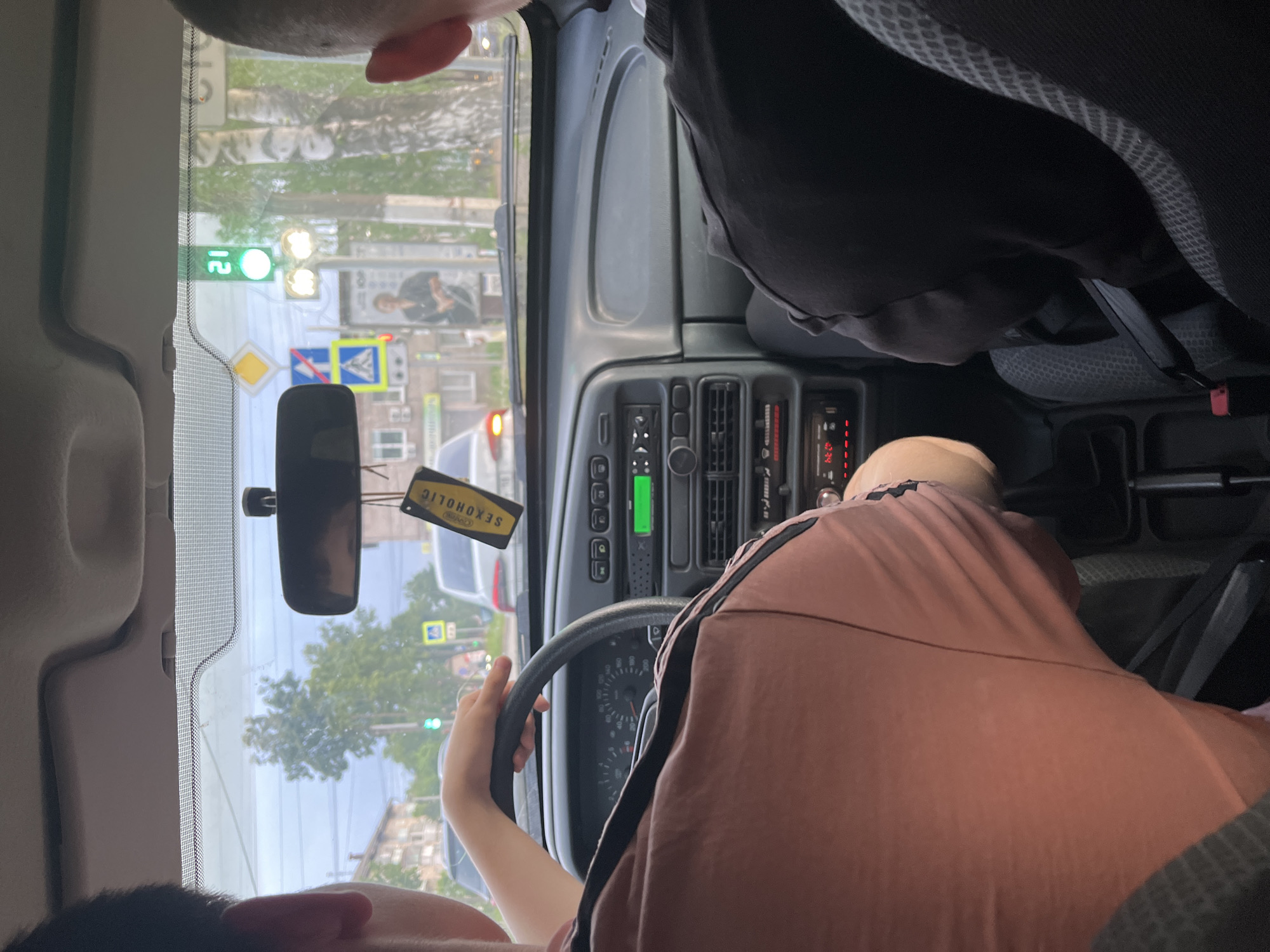 The image shows an interior view from a car in Chaikovsky, Russia. A person with light skin is driving, wearing a pink shirt with dark stripes, holding the steering wheel. The dashboard has a green display and various buttons and vents. A rearview mirror hangs with a yellow air freshener displaying the word "SEXOHOLIC" in reverse. Outside the windshield, traffic lights are visible showing a green light. There is a street sign with a photograph of a man on it and other signs with traffic information, including one indicating a pedestrian crossing. The city environment includes buildings and trees. Another person can be seen in the passenger seat wearing a dark colored shirt. The camera angle is from the back seat.