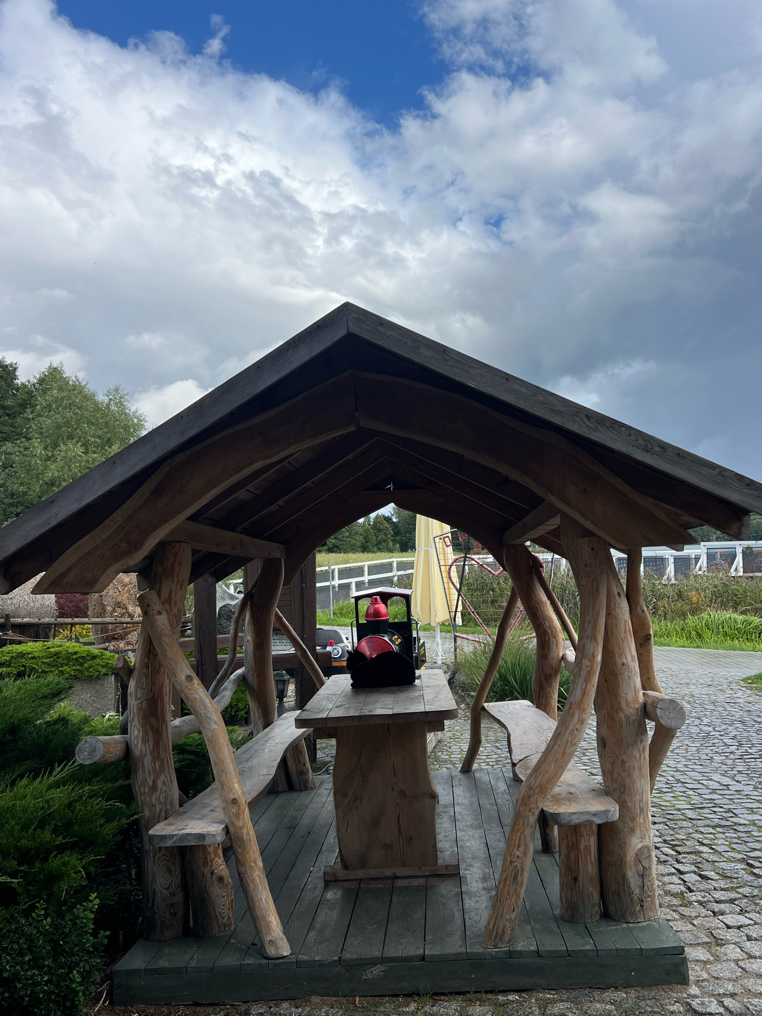 A wooden gazebo with a rustic design stands on a paved area in Mrągowo, Poland. The gazebo has a pitched roof supported by rough-hewn wooden posts. Inside, a wooden picnic table and benches are visible. On the table, there is a black and red children's toy train. The area surrounding the gazebo is covered in cobblestones, with greenery and a white fence in the background. The sky above is cloudy, with patches of blue.