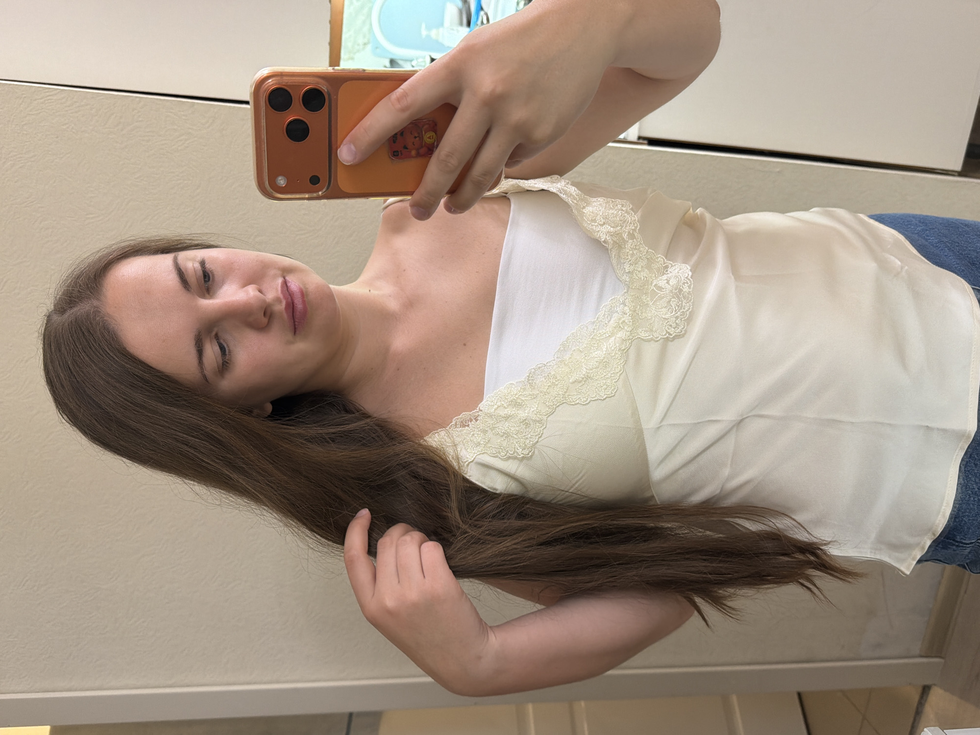 A young woman is taking a selfie with a mobile phone. She is wearing a white tank top with lace trim over a white top and blue jeans. She is holding a strand of her long, brown hair with her left hand. The background appears to be an interior setting, possibly a room with white cabinets and walls. There is no information to determine that it is located in Sochi, Russia.