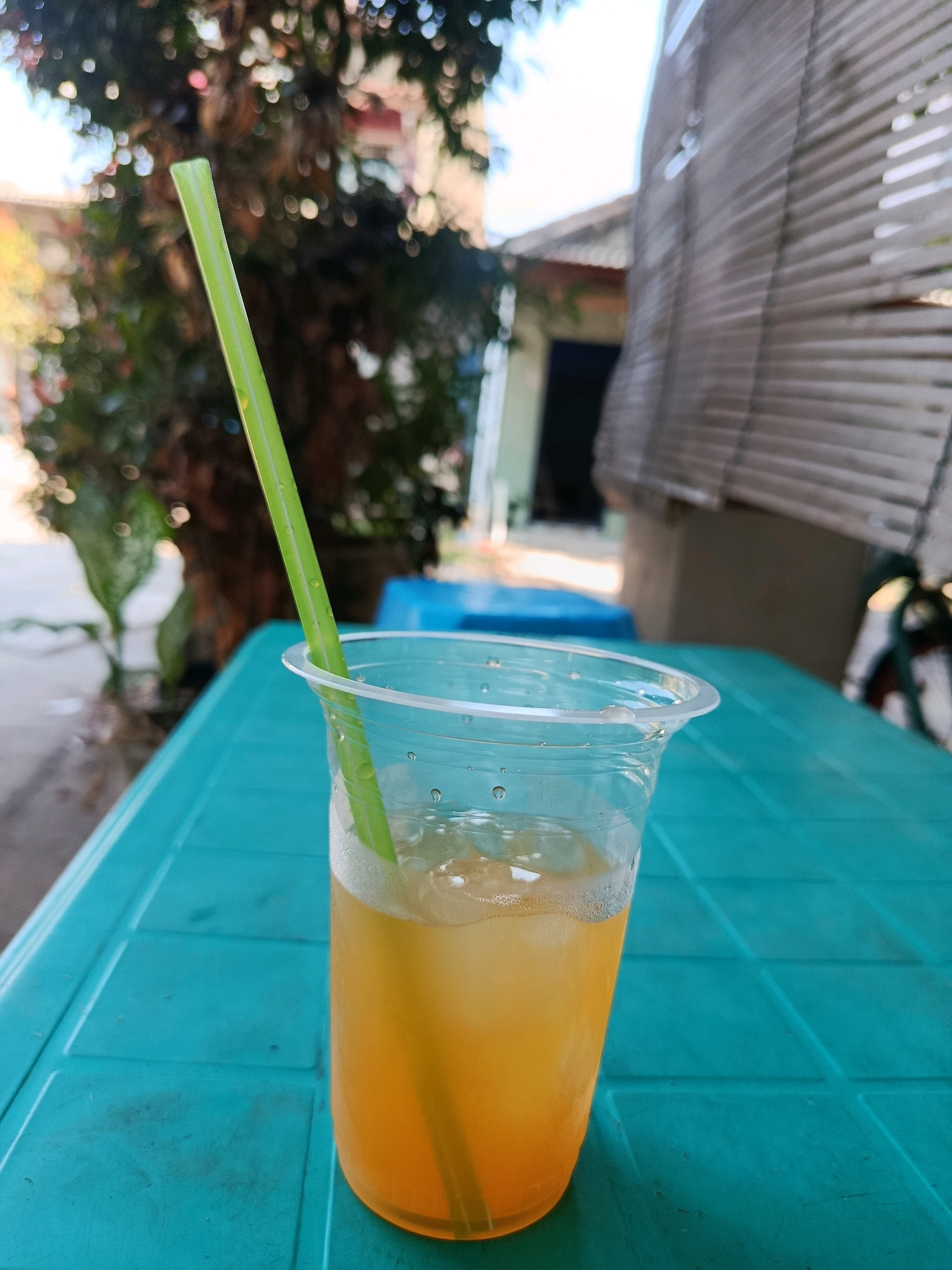A close-up, eye-level shot shows a clear plastic cup filled with an orange beverage and ice cubes, with a green straw inserted. The cup rests on a turquoise table. In the background, out of focus, is a tree with green leaves and a building with a shuttered window. The scene appears to be an outdoor setting in Jobokuto, Indonesia.
