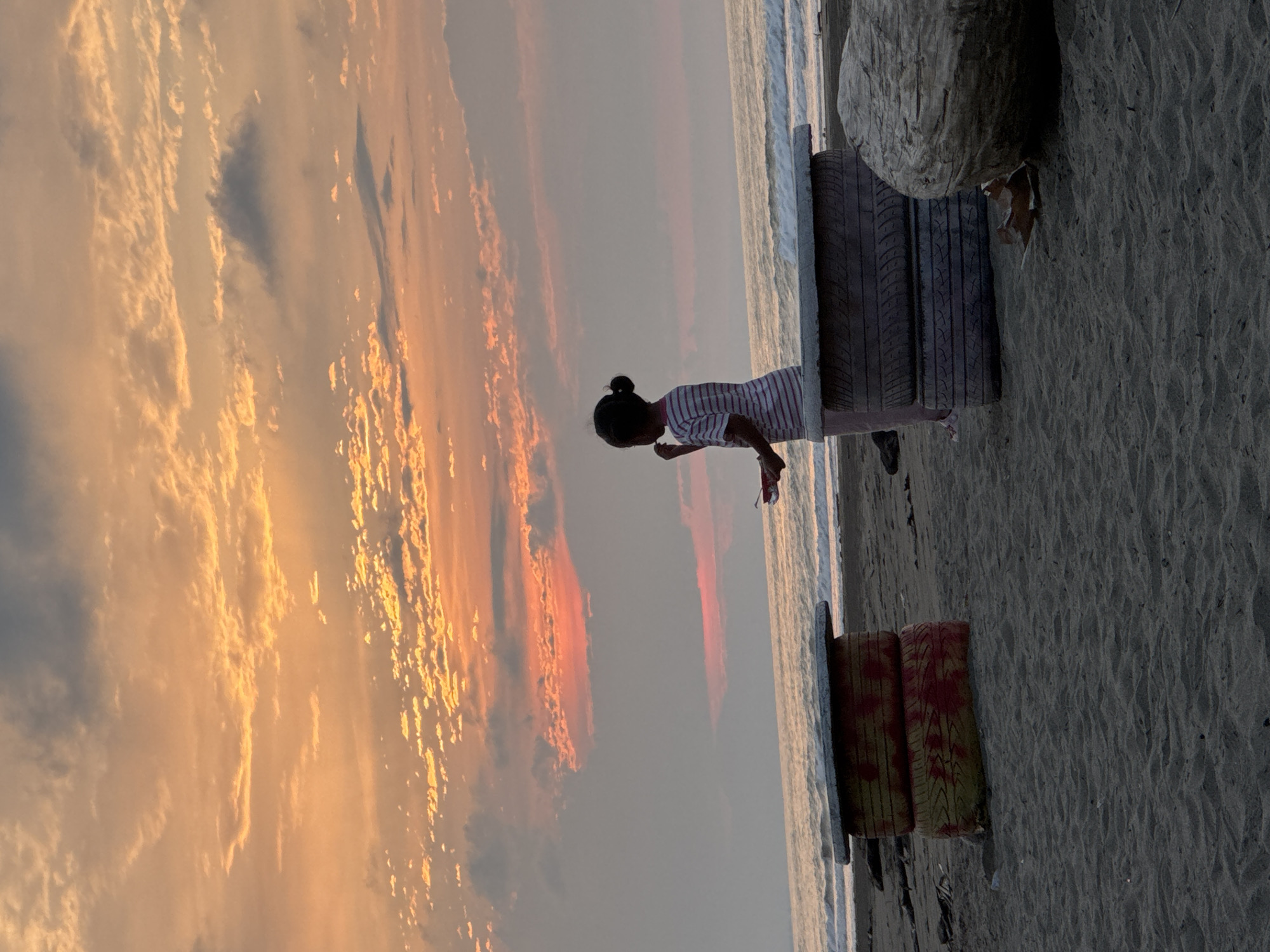 A young child stands on a sandy beach in Reudeuep, Indonesia, facing the ocean at dusk. The sky is a vibrant mix of orange, pink, and grey clouds, illuminated by the setting sun. The child is silhouetted against the colorful sky, wearing a striped shirt and light-colored pants. They are holding something small and red in their right hand.
In the foreground, two makeshift tables or benches are visible. The one on the left is constructed from stacked, painted tires, with visible red and yellow markings. The table on the right is similarly constructed from tires, with a flat concrete top. A large piece of driftwood lies on the sand to the right of the right-hand table. The ocean waves gently roll onto the shore in the background. The overall scene depicts a peaceful moment at the beach as the day ends.