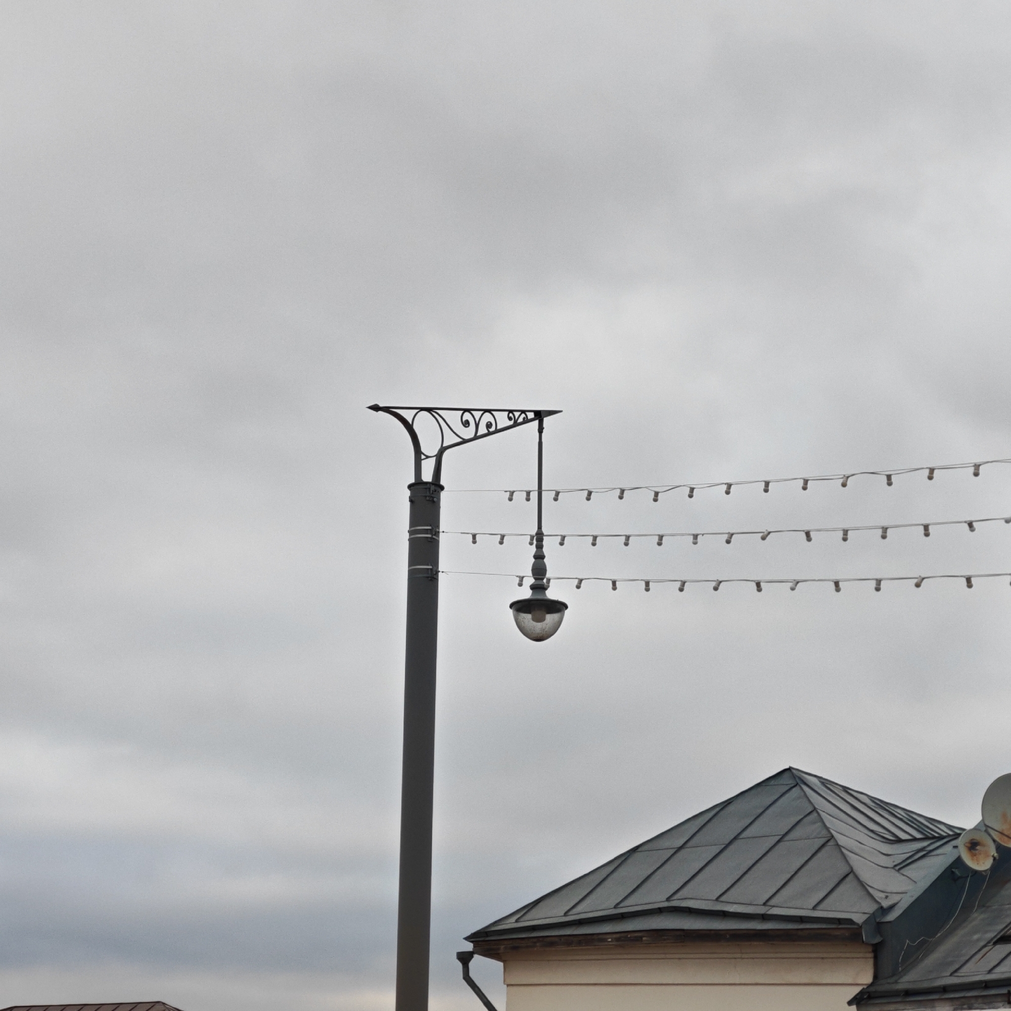 The image presents a low-angle view of a street scene, likely in an urban setting in Rostov, Russia, characterized by overcast skies. A prominent feature is a gray, ornate street lamp post. Attached to the lamp post are three strands of small decorative lights. Part of a building is visible in the lower right corner of the frame, its roof made of gray metal and includes a rain gutter. Two satellite dishes are attached to the roof. The building's facade is a pale, neutral color. The general atmosphere is muted due to the overcast sky.