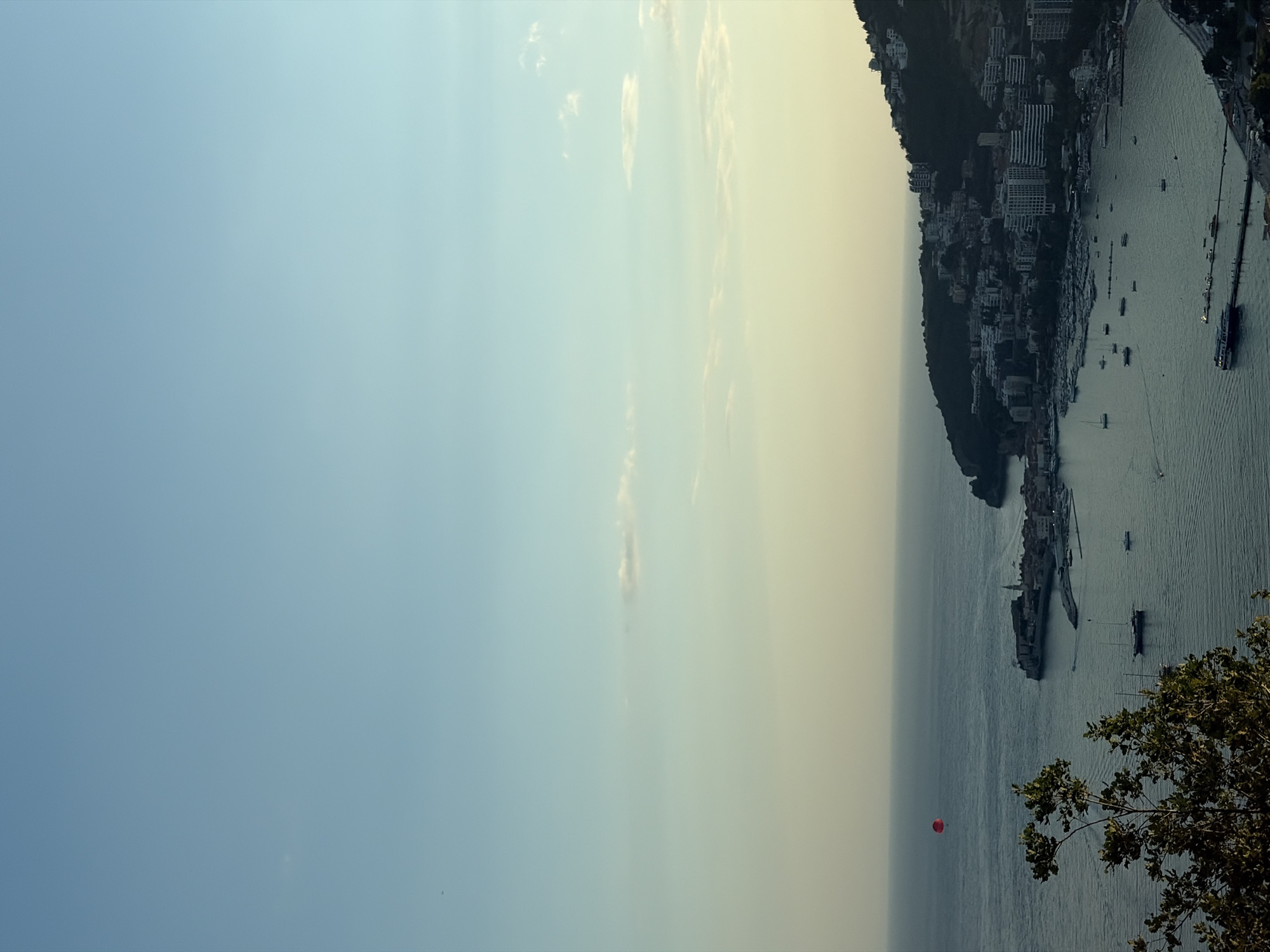 This is a vertical, high-angle shot of a coastal town nestled on a hillside overlooking a bay. The sky transitions from a clear, light blue on the left to a hazy, yellow-tinged sky on the right, suggesting either sunrise or sunset. Scattered clouds dot the sky. The bay is filled with numerous boats, appearing to be moored or anchored, with a larger vessel docked along a pier. The town itself is densely packed with buildings, a mix of white and darker structures, climbing up the steep terrain. Lush green trees are visible on the hillside and in the foreground, on the right side of the frame. In the middle distance, over the water, a small red balloon is visible, floating in the air. The overall scene evokes a serene and picturesque coastal landscape.