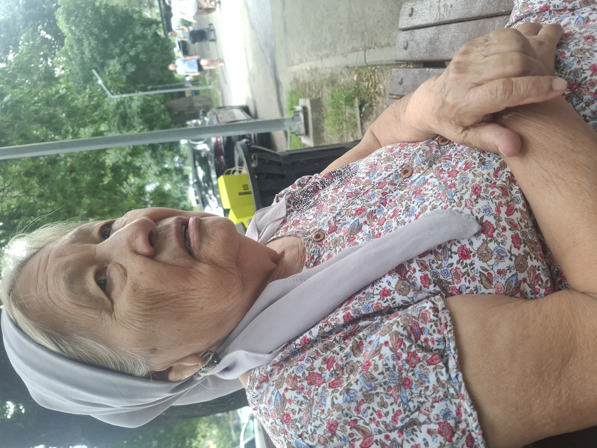 An elderly woman with fair skin and white hair, wearing a light gray headscarf and a floral print dress, sits on a wooden bench outdoors. She is looking to her right with her mouth slightly open. Her hands are clasped in her lap. Behind her, a sidewalk runs alongside a grassy area with trees and a lamppost. In the background, cars are parked, and several people are walking, one pushing a stroller. The scene appears to be in a public park or street in Almaty, Kazakhstan.