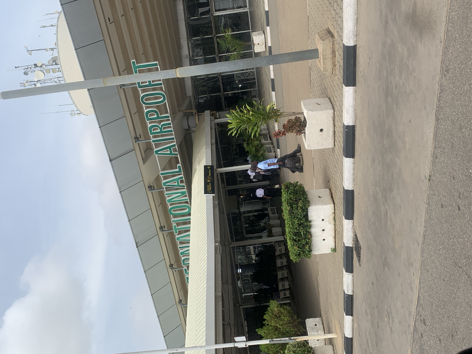 The image captures the entrance area of an airport building, identified by the prominent green lettering spelling "INTERNATIONAL AIRPORT" on its facade. The building features a modern design with large glass windows and a wide canopy over the entrance.
Below the main sign, a smaller white sign with arrows indicates "Domestic Departure" and "International Departure." Several individuals are visible near the entrance, suggesting typical airport activity. One person in a light blue shirt and dark trousers is standing next to a concrete planter, carrying a black backpack. Other people in white shirts can be seen further inside the entrance.
The foreground shows a paved area, likely a pedestrian walkway or drop-off zone, bordered by a black and white striped curb. Concrete planters filled with green shrubs and smaller plants are positioned along the building's edge. A plain grey pole, possibly a signpost or light post, stands vertically in the mid-ground.
Above the airport building, against a partly cloudy sky, a collection of antennas and satellite dishes is visible on a rooftop or tower, indicative of communication infrastructure common at airports. The scene is brightly lit, suggesting daytime conditions. The setting is an international airport in Gwaska, Nigeria.