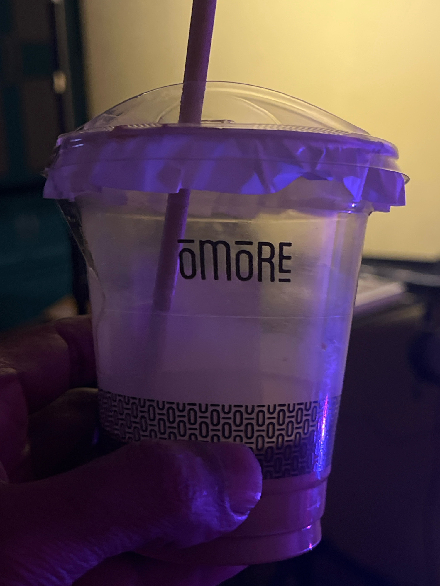 A plastic cup with a clear lid and a straw is held in a hand, illuminated by purple and yellow light. The cup has a logo that reads "TOMORE" in black lettering and a patterned band near the bottom. The background is blurred, showing a wall and some objects in the distance. The image appears to be taken indoors, possibly in a cafe or restaurant.
