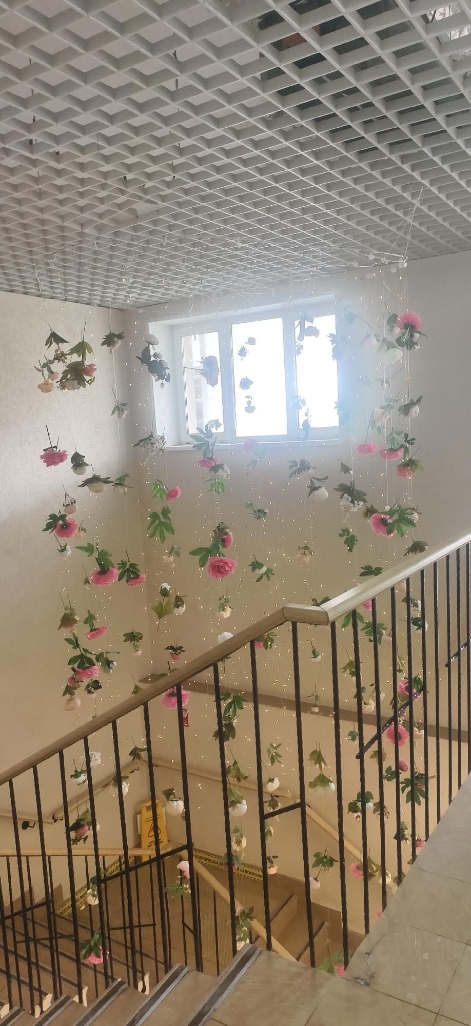 An indoor scene showcases a brightly lit staircase adorned with numerous hanging artificial flowers and fairy lights, creating a festive atmosphere. The perspective is from the top of the stairs, looking down and across.
The ceiling features a white grid-panel design, from which thin lines suspend a multitude of artificial flowers. These flowers are predominantly pink and white, interspersed with green leaves, and appear to be of various sizes and types, suggesting peonies or roses. Strands of small, warm-toned fairy lights are woven amongst the hanging floral arrangements, adding a sparkling effect.
In the background, a large, multi-paned window with a white frame allows bright daylight to flood the space, causing a slight glare. The walls are a light, neutral color.
The staircase itself has dark metal railings with a lighter wooden handrail. The steps are constructed from light-colored tiles, each featuring a darker anti-slip strip. Below, on a landing, a yellow "wet floor" sign is partially visible. Additionally, white tape with Russian text can be seen on the wall or railing, which reads, "ПОЖАЛУЙСТА МОЙТЕ" (Please wash) and "НЕ ВЫХОДИТЬ ЗА БОРТИК" (Do not go over the edge/side), though some letters are obscured. No people are present, and the time of day appears to be daytime due to the bright light from the window. The decorations suggest a celebratory event or a permanent decorative enhancement within what appears to be a public or semi-public building in Votkinsk, Russia.